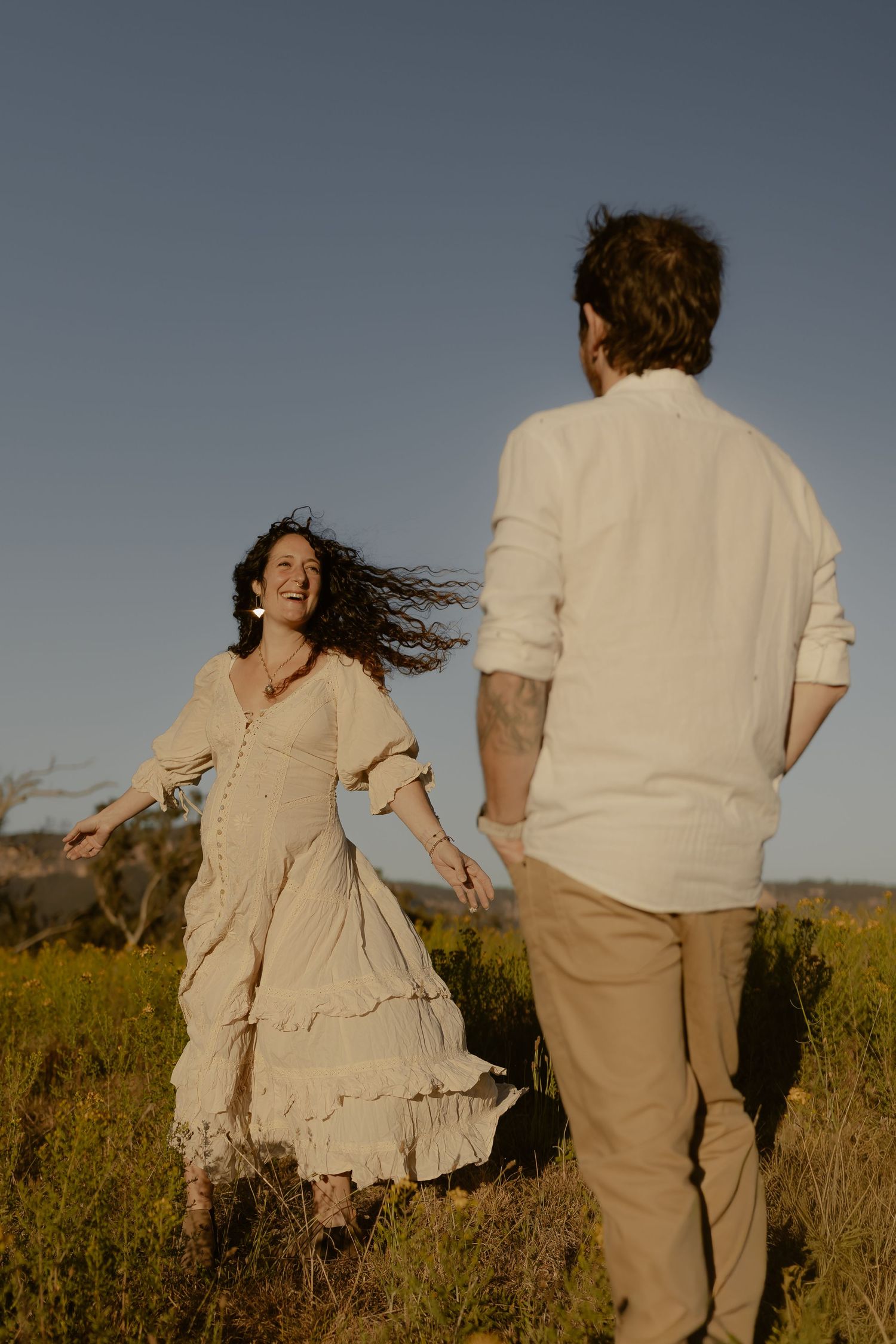 A couple in light colored clothing runs playfully through a golden field at sunset.