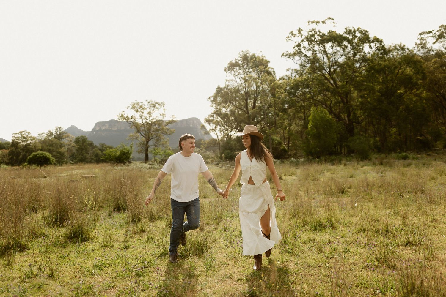A couple runs hand in hand through a grassy field with mountains visible in the distance.