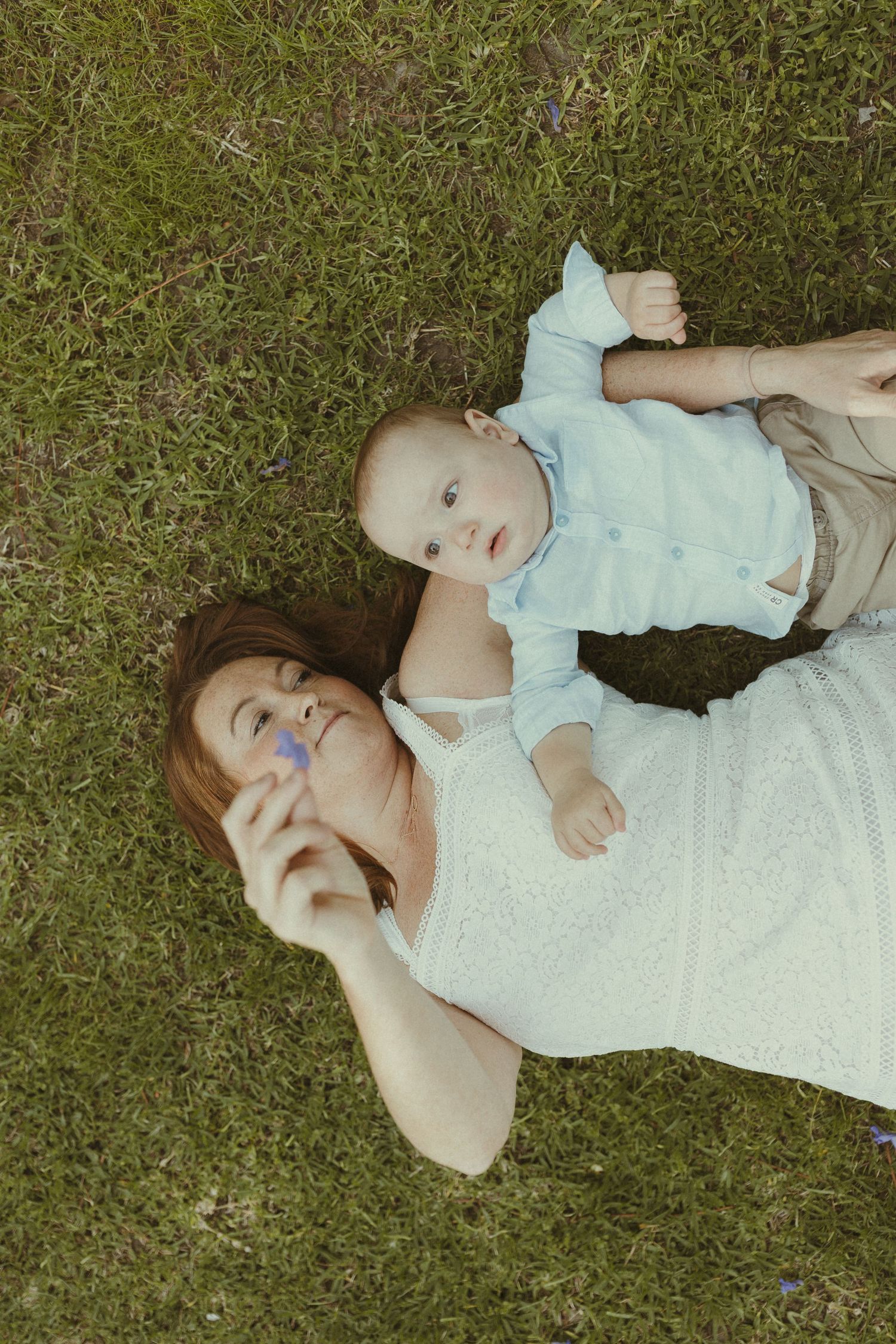 Two people lying on green grass wearing light colored clothing on a sunny day.