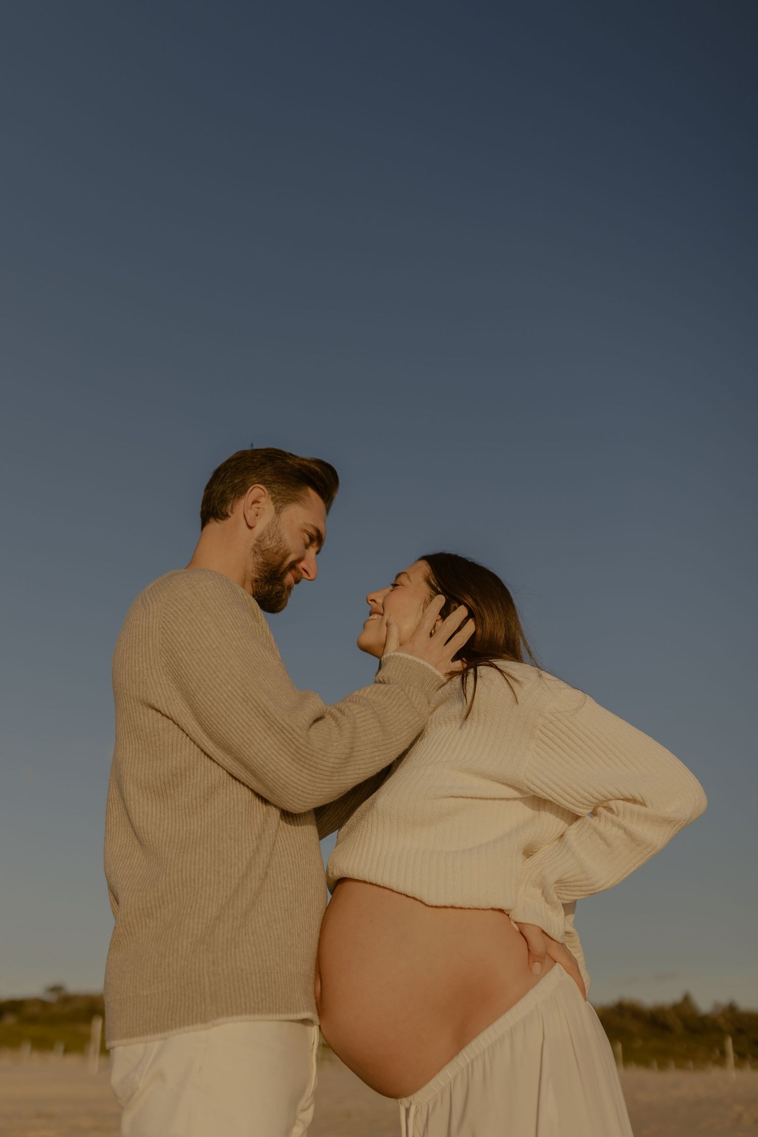 Intimate maternity photoshoot on beach with couple in neutral sweaters at dusk.