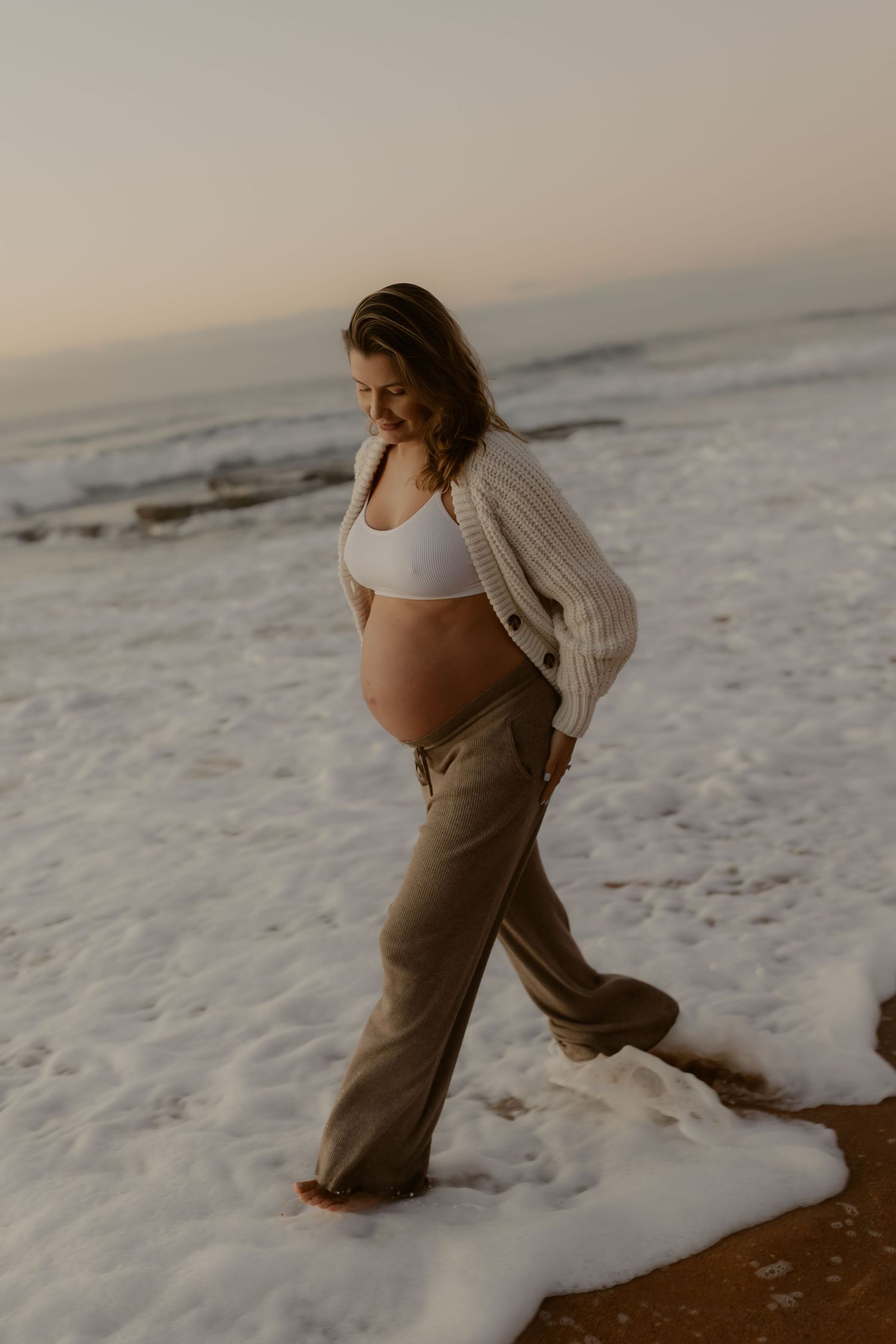 Pregnant woman in white crop top and khaki pants walking in ocean waves at sunset.
