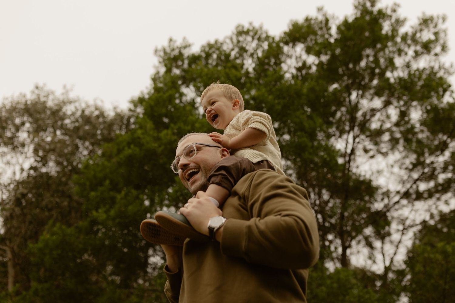 Father smiling with laughing toddler sitting on his shoulders outdoors among green trees.