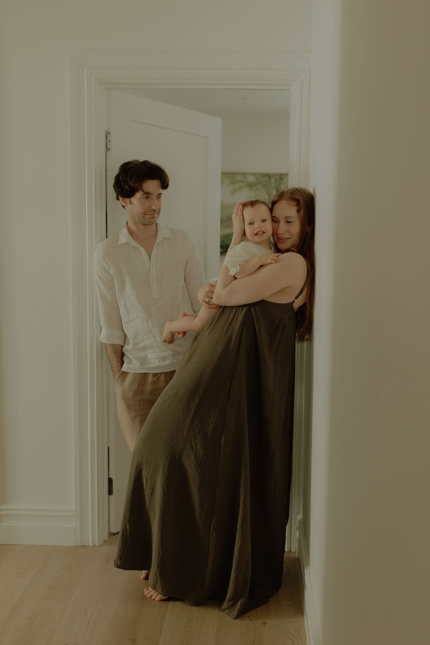 A young family with a mother in olive dress holding a baby, standing in a bright doorway with a man in white shirt.