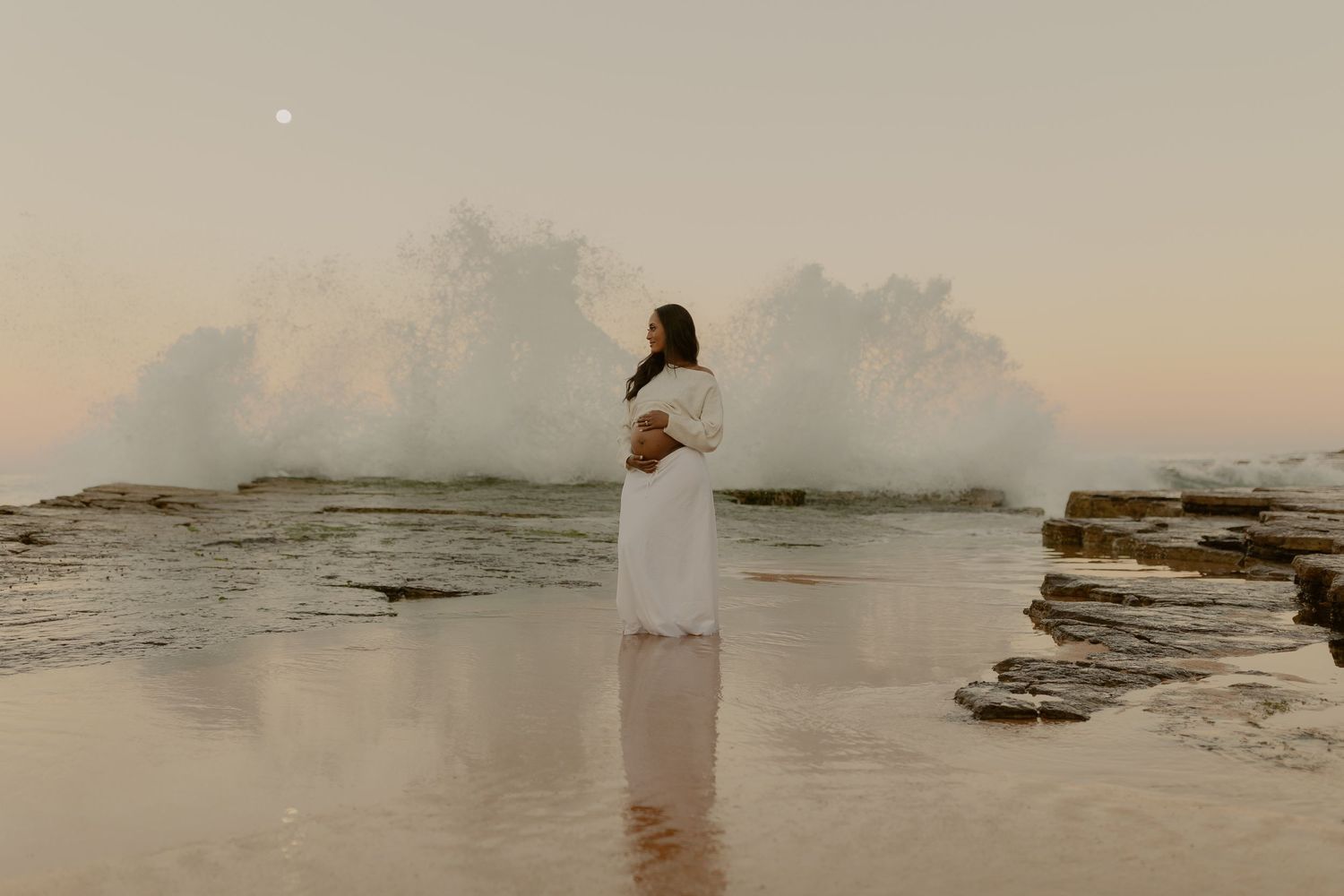 Pregnant woman in white dress standing on rocky shore with dramatic crashing wave at sunset.
