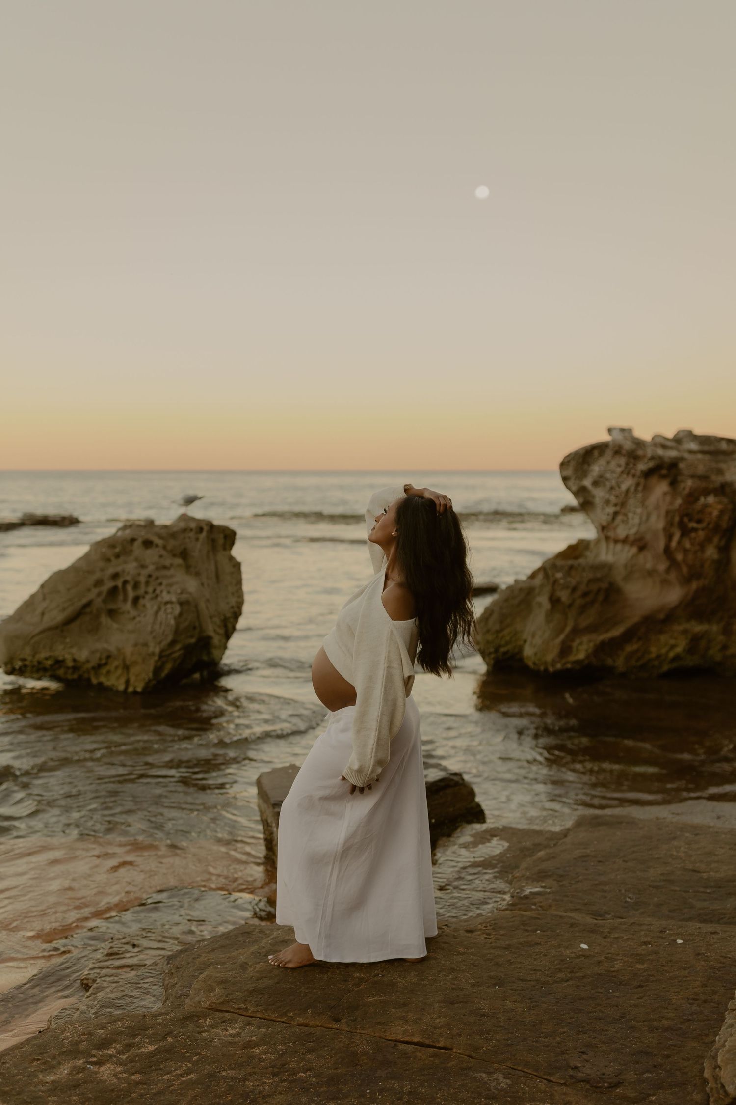 Woman in white dress sitting on rocks by the ocean at sunset with moon visible in sky.