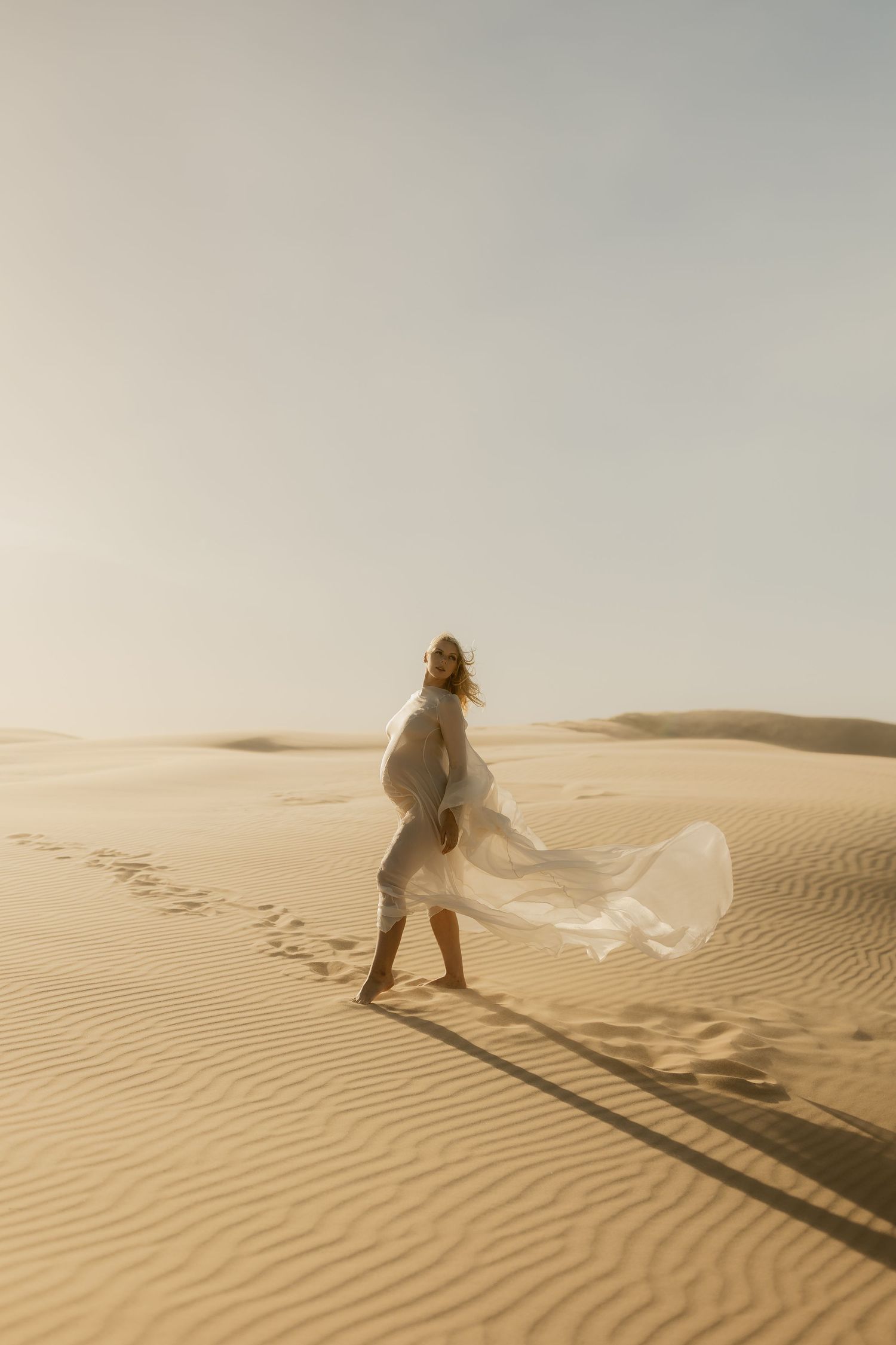 Woman in flowing white dress walking on golden sand dunes in desert sunlight.