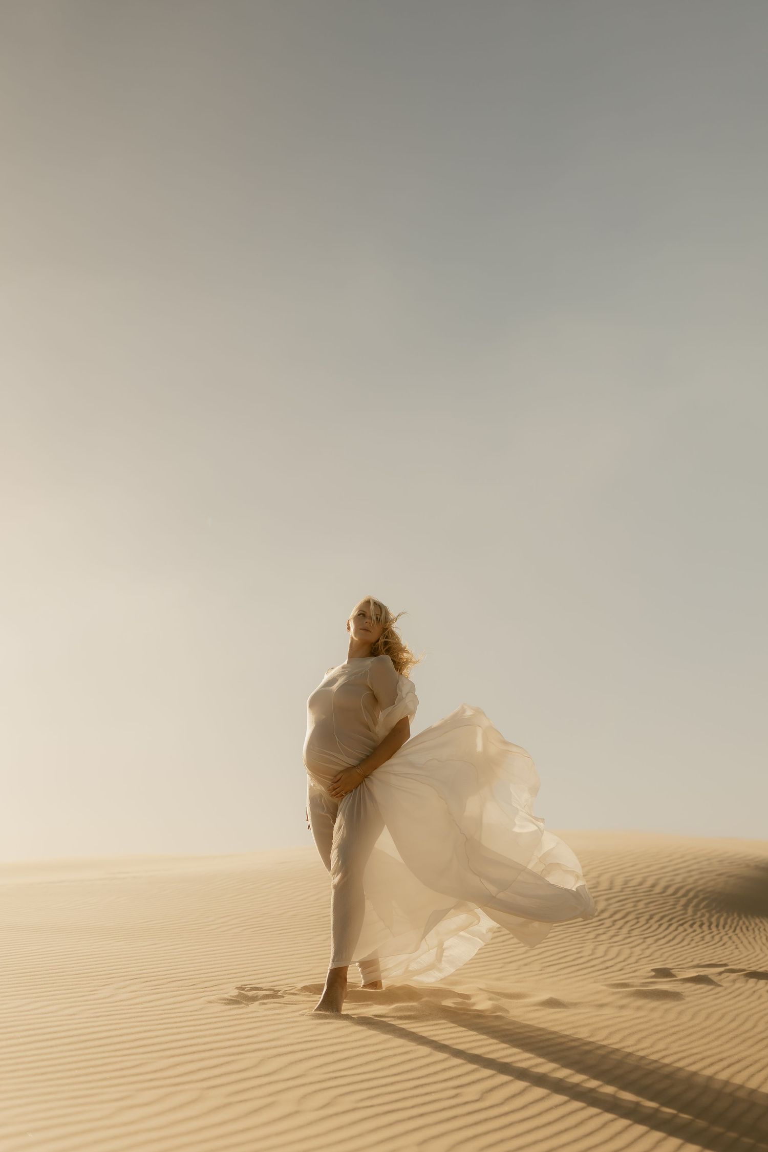 Pregnant woman in flowing white dress walking on desert sand dunes at golden hour.