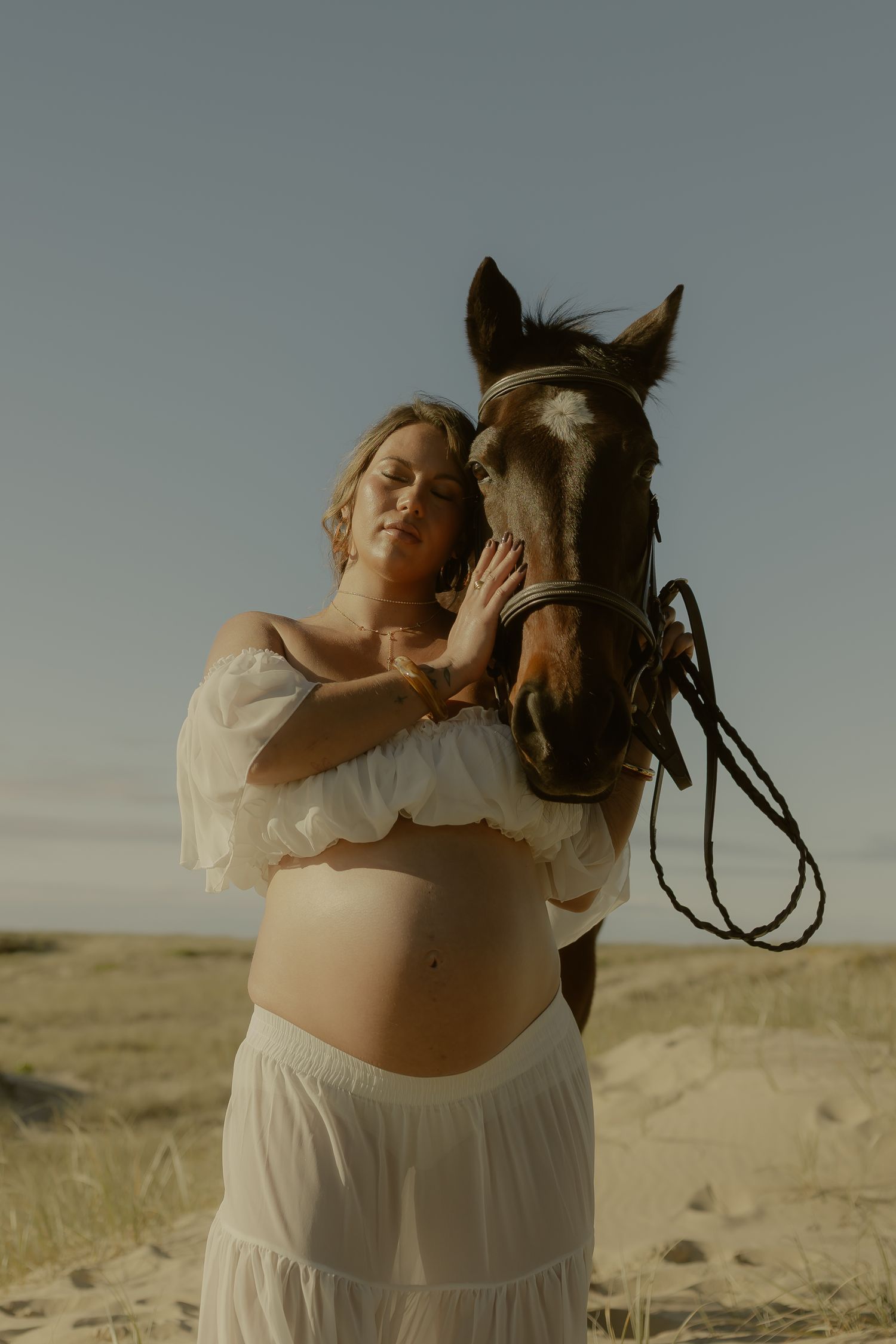 Pregnant woman in white outfit tenderly touches a brown horse's face on a sandy landscape.