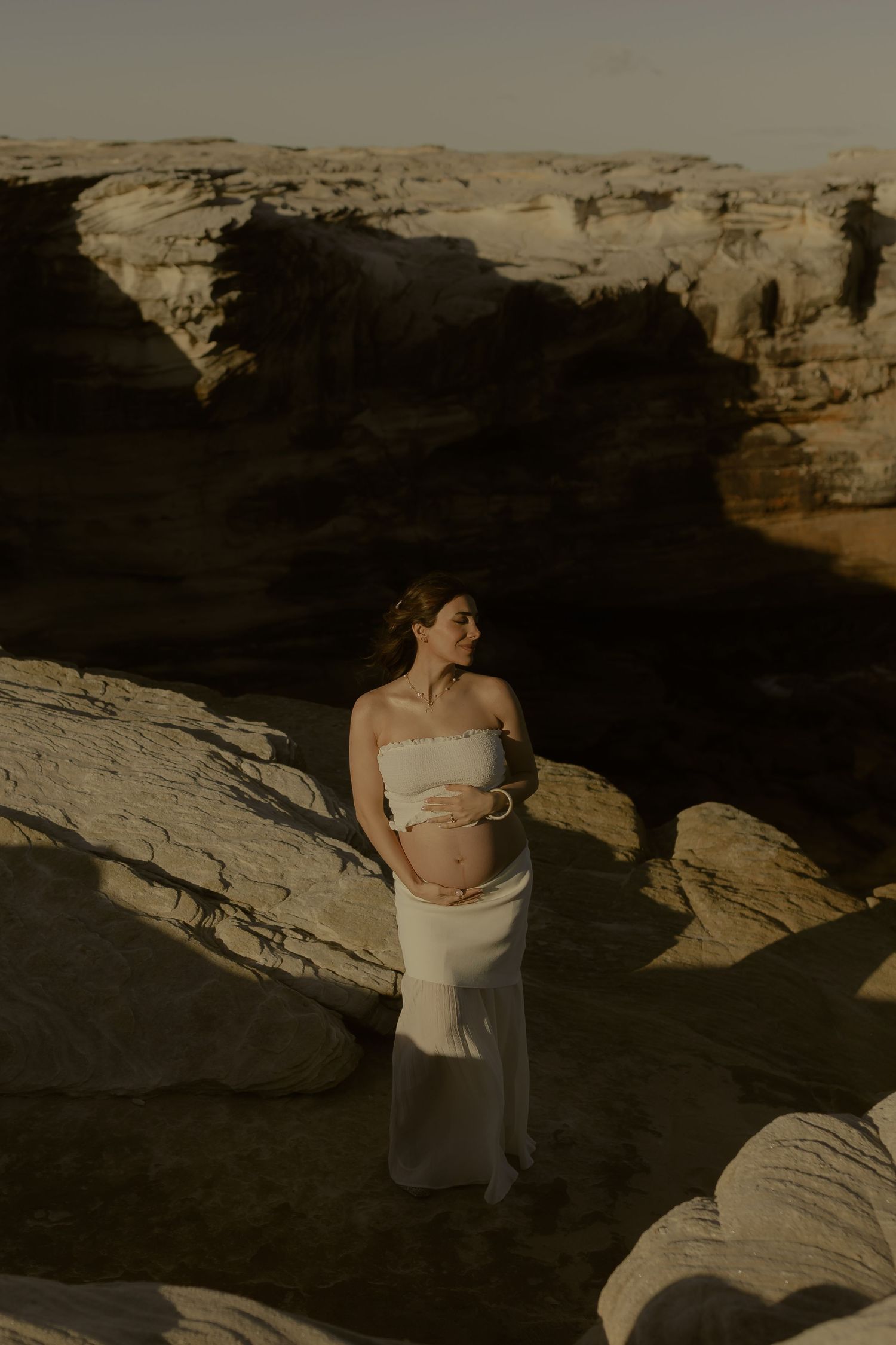 Pregnant woman in white crop top and skirt cradling her baby bump among dramatic desert canyon rock formations.
