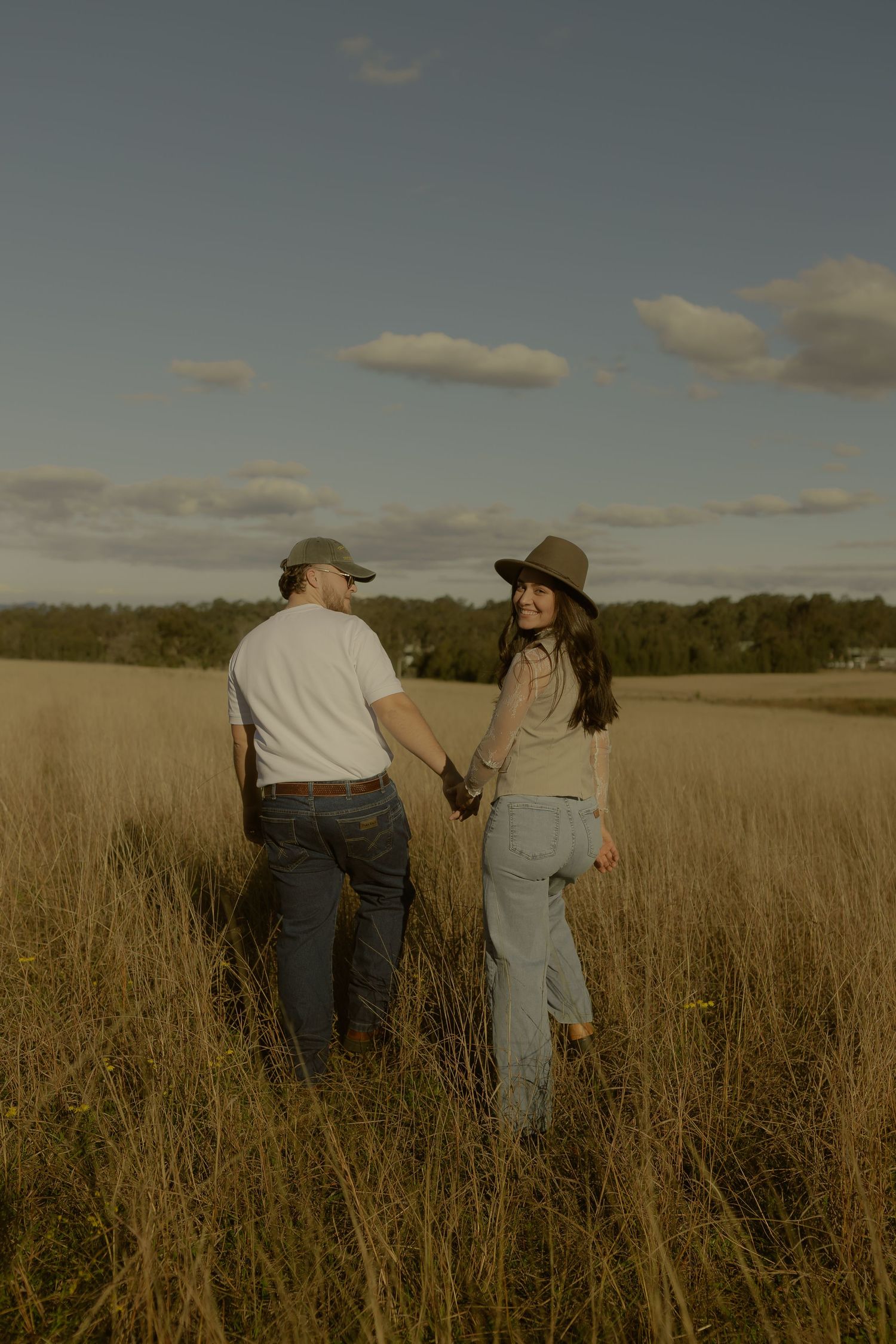 Couple holding hands walking through golden wheat field under blue sky with scattered clouds.