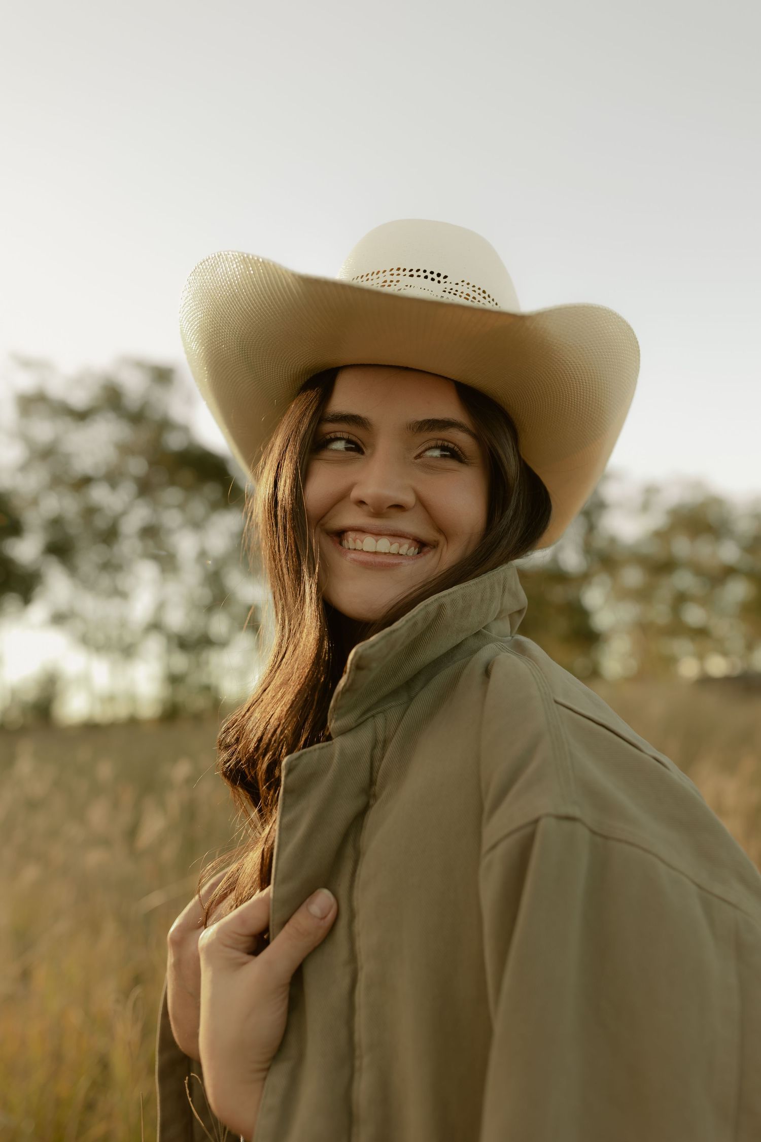 Smiling woman in cowboy hat and olive jacket poses outdoors in golden field at sunset.