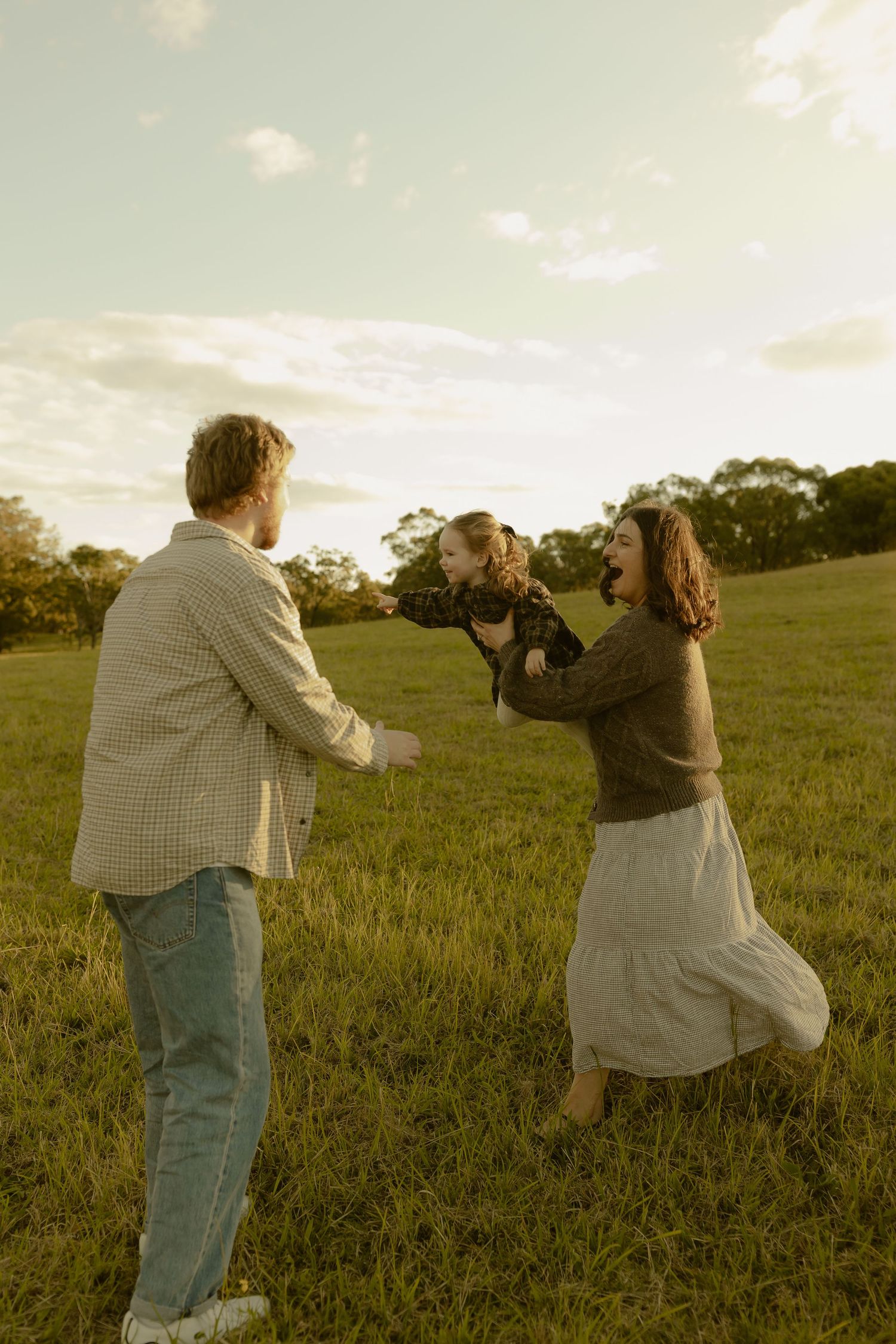 Family playing in green field at golden hour, parents lifting child with joy.