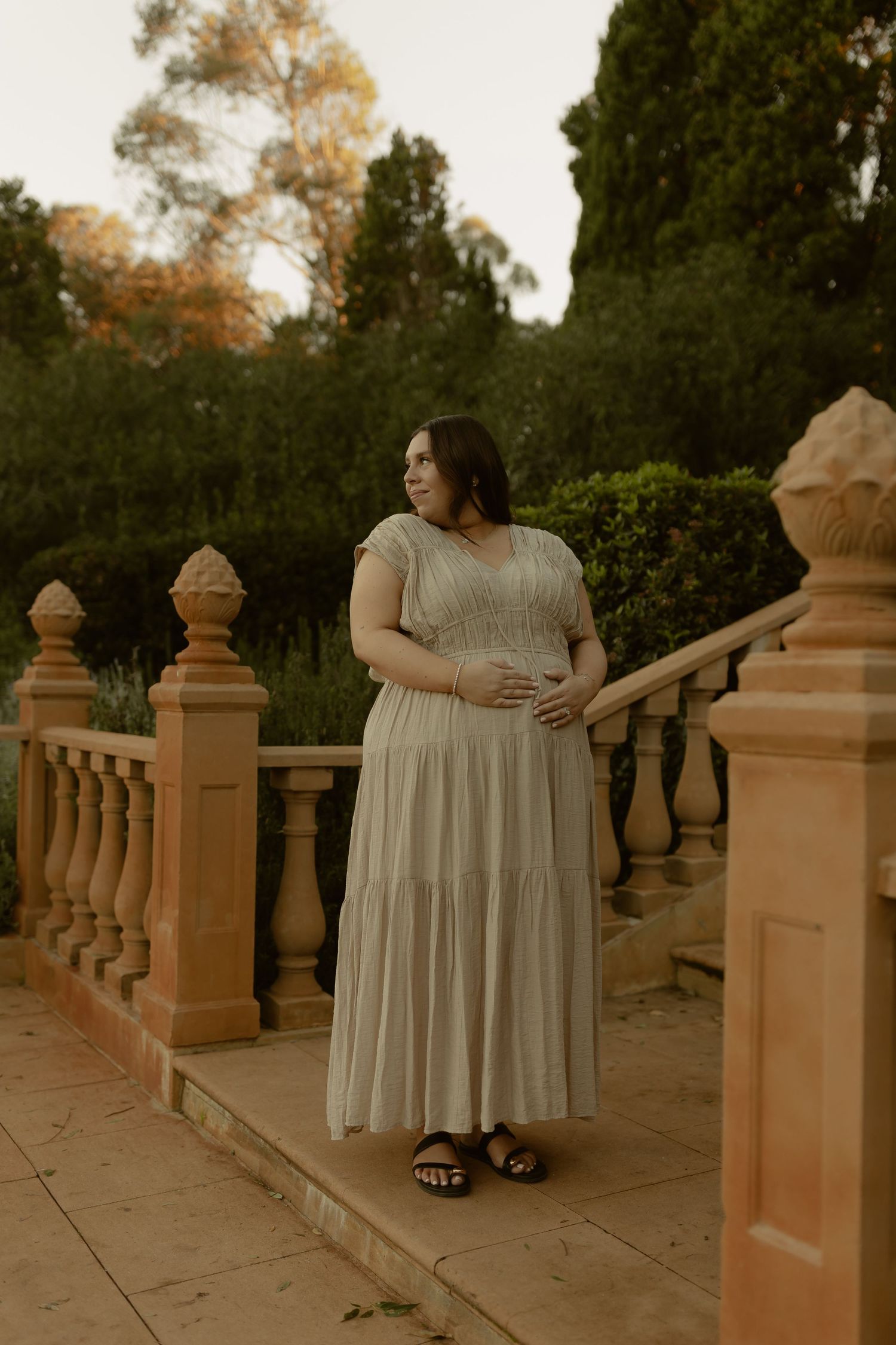 Pregnant woman in sage green maxi dress stands on ornate stone balustrade in lush garden at golden hour.