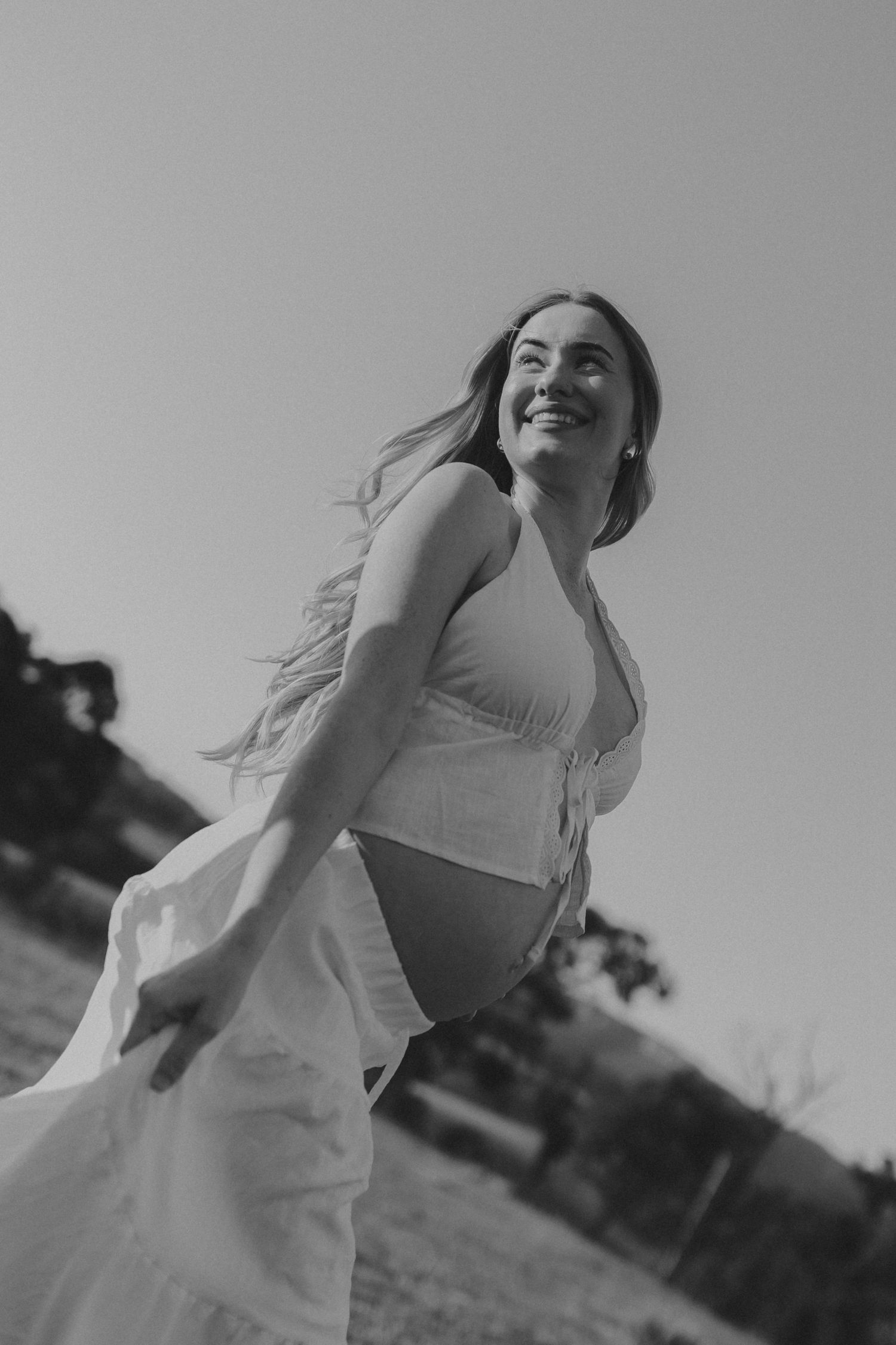 Black and white photo of a smiling woman in a white dress, hair flowing in the wind outdoors.