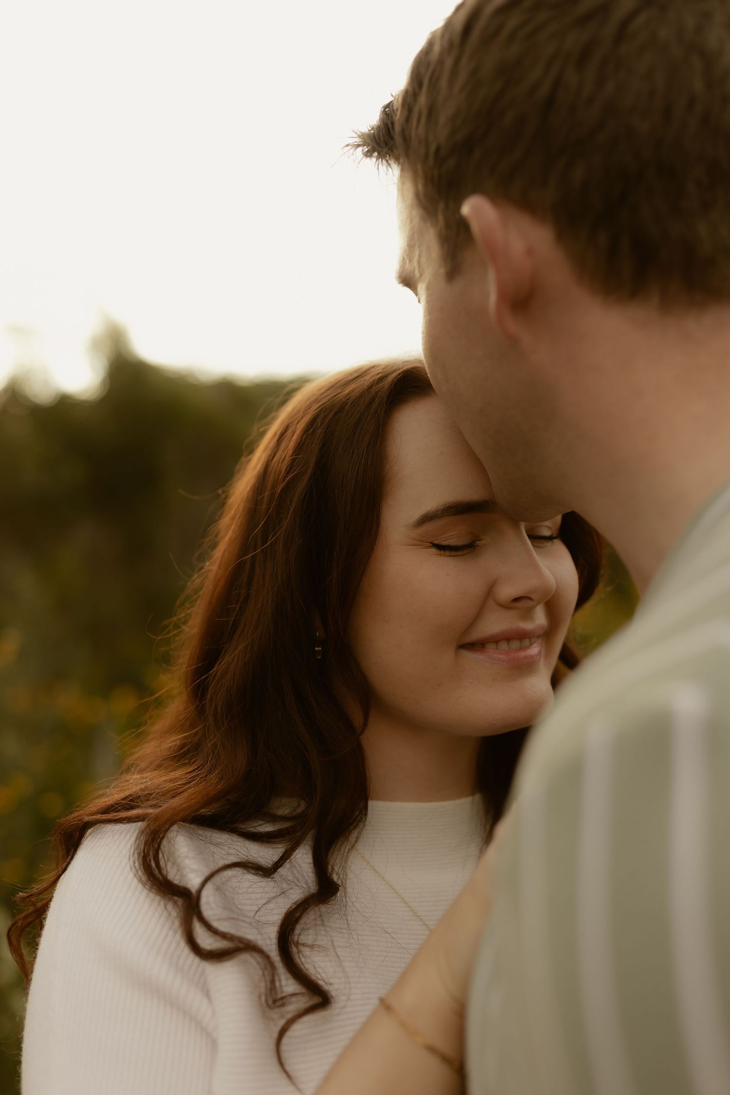 A smiling woman with auburn hair closes her eyes as she leans toward a man outdoors at golden hour.