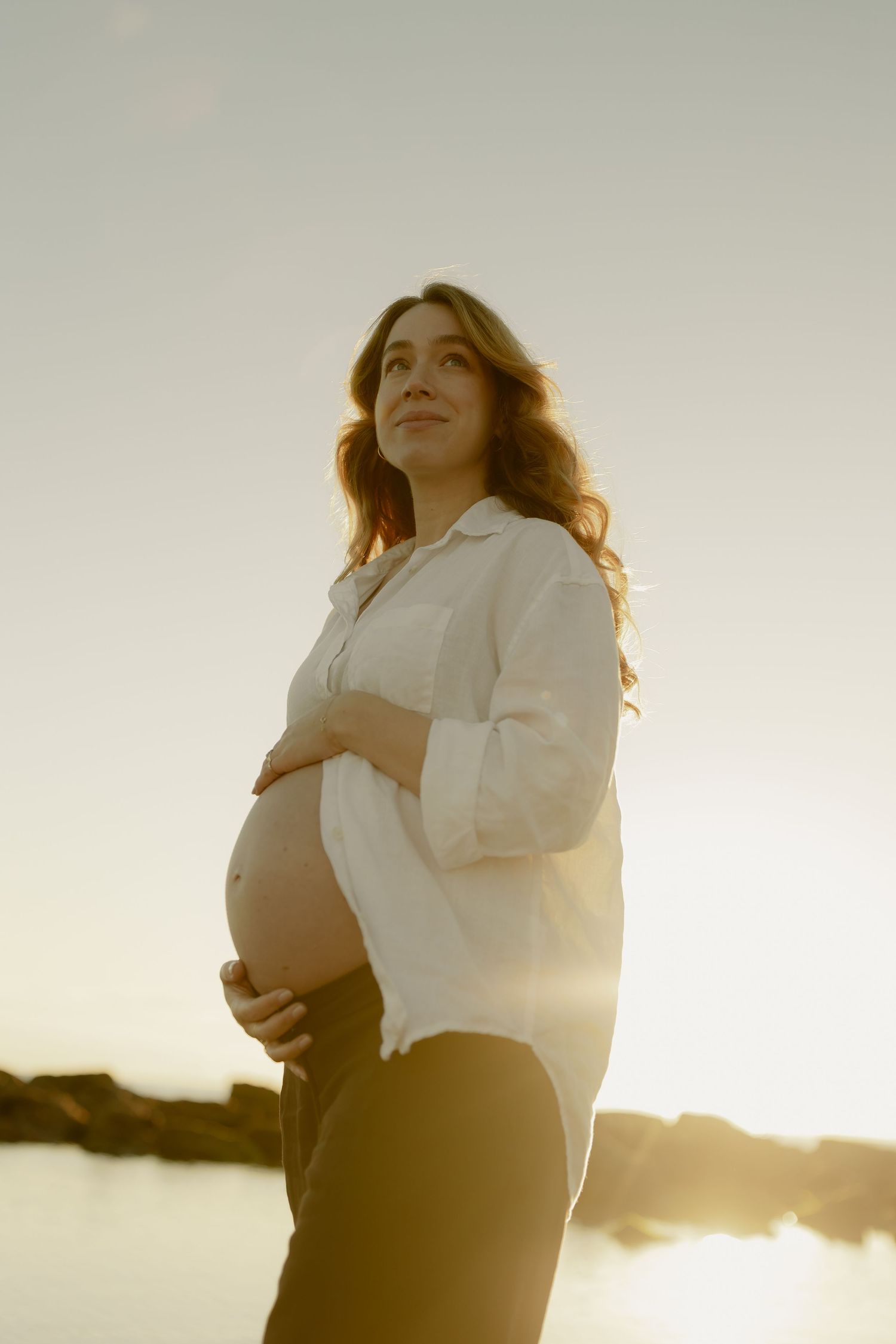 Pregnant woman in white linen shirt cradles bump on beach, gazing upward in warm sunset glow.