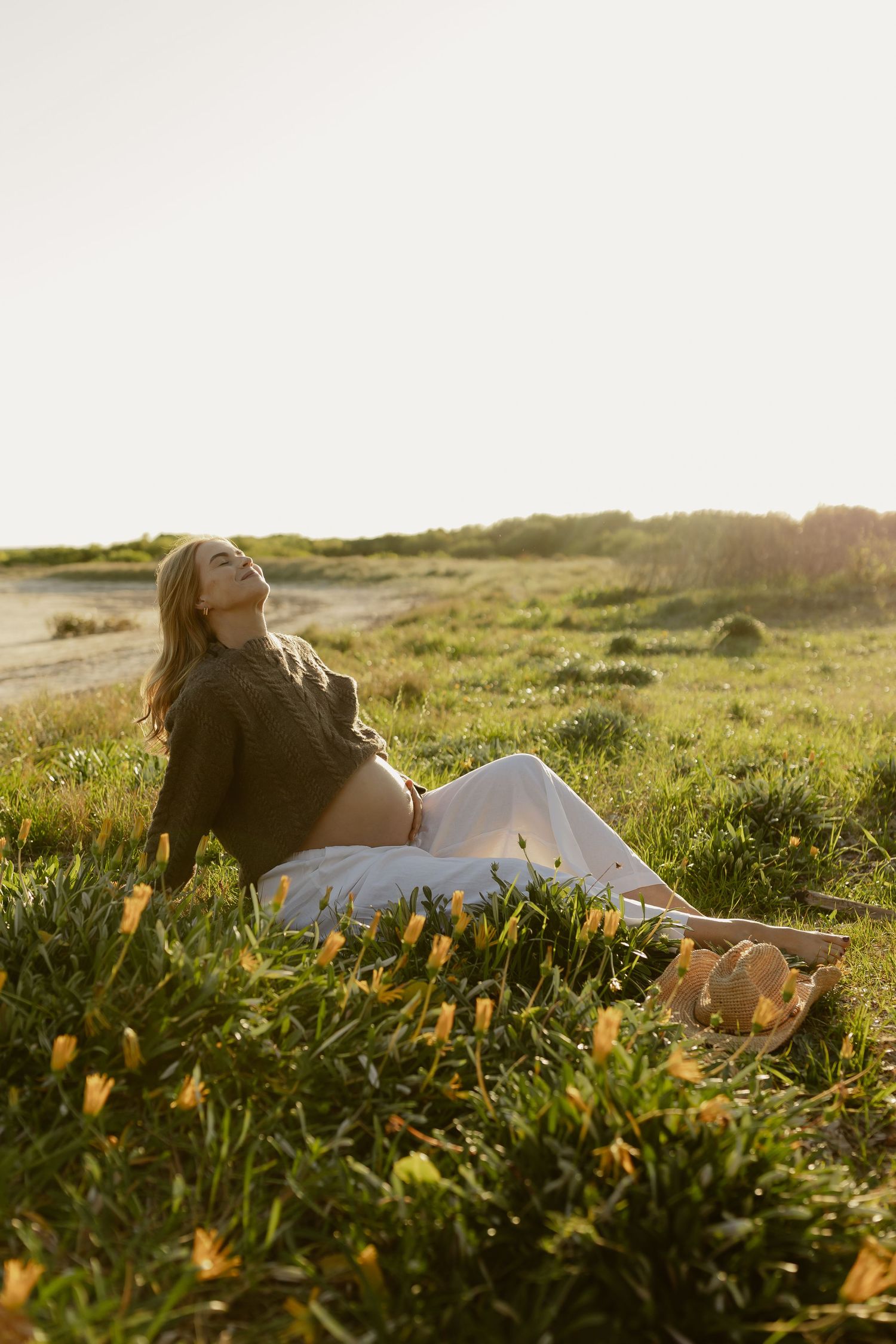 Pregnant woman sitting in a sunny green meadow with yellow wildflowers, wearing a green jacket and white pants.
