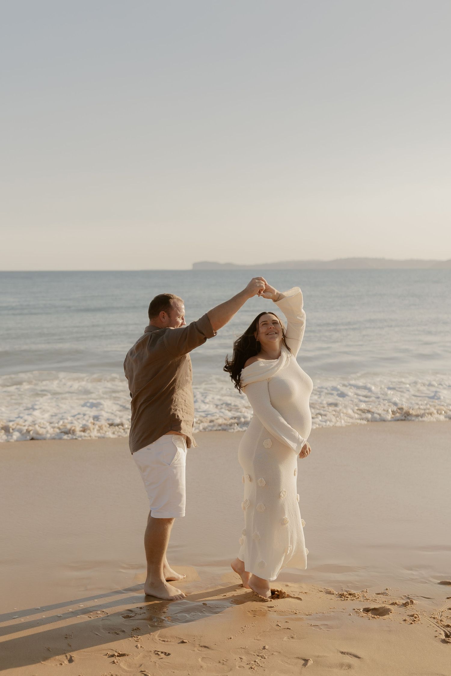 A couple in white outfits dances together on a sandy beach during golden hour.