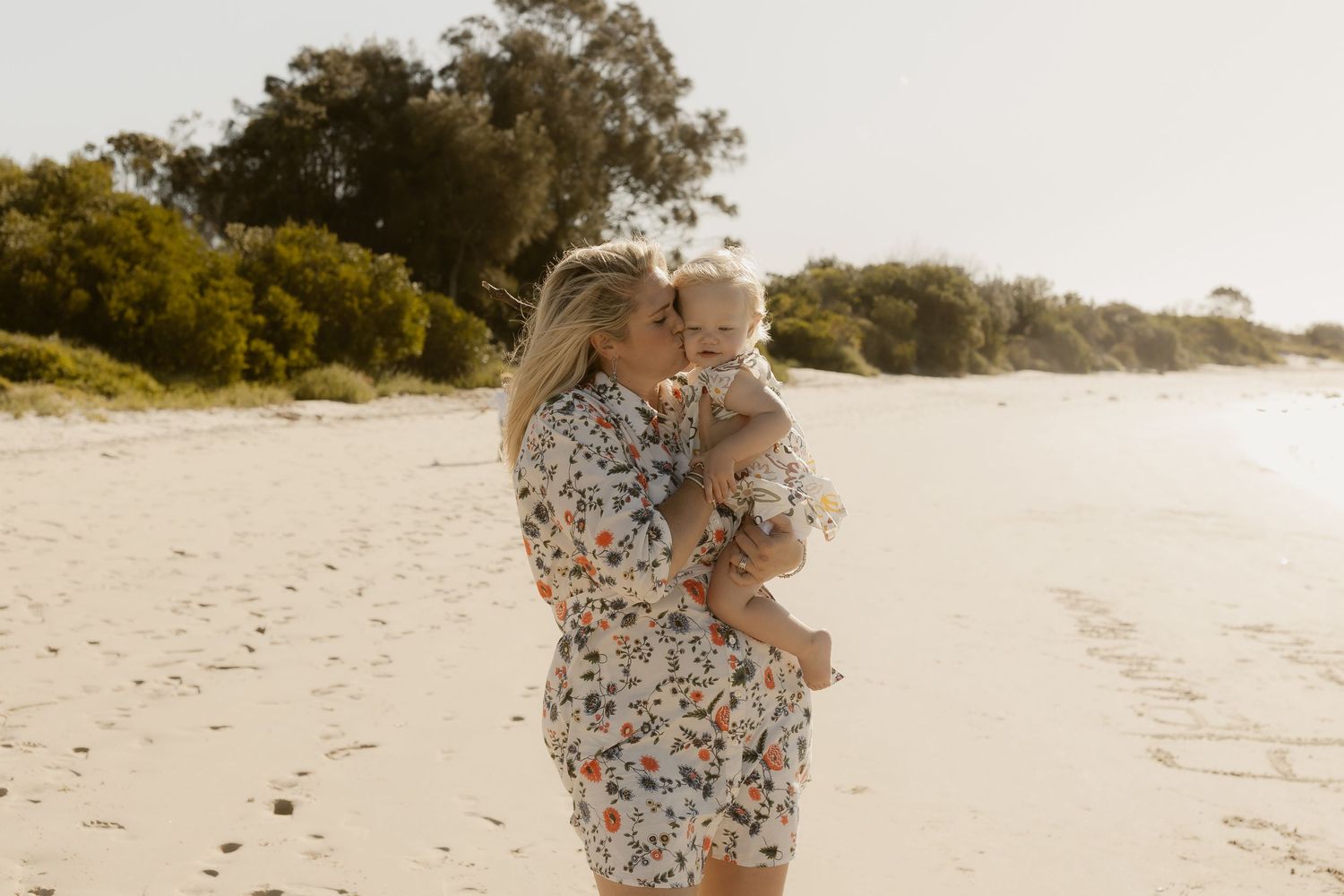 A tender embrace between parent and child on a sunny beach with trees in the background.