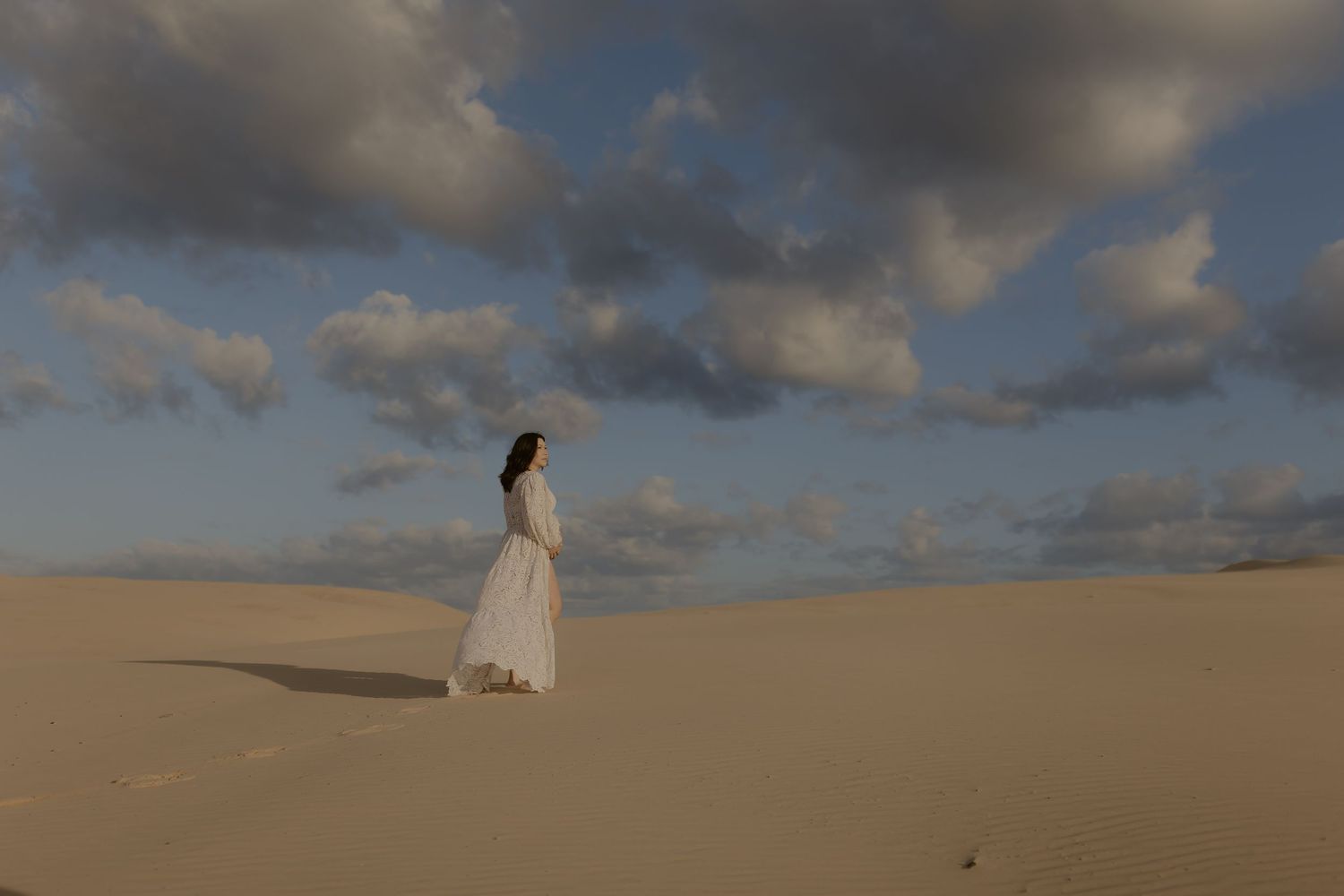 A person in a flowing white dress walks across golden sand dunes under a dramatic cloudy sky at sunset.