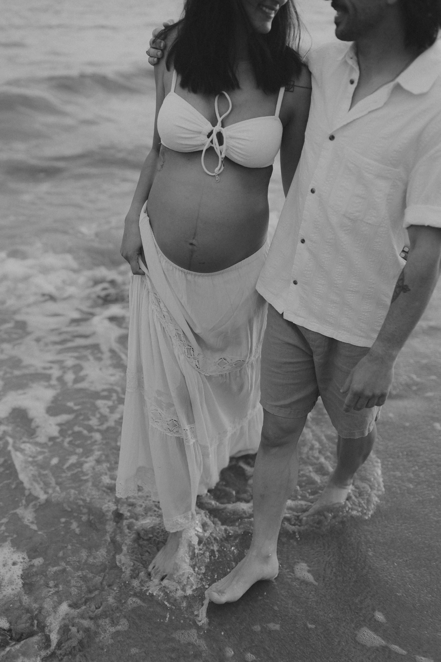 A couple's legs standing in ocean waves, with her wearing a flowing white dress.