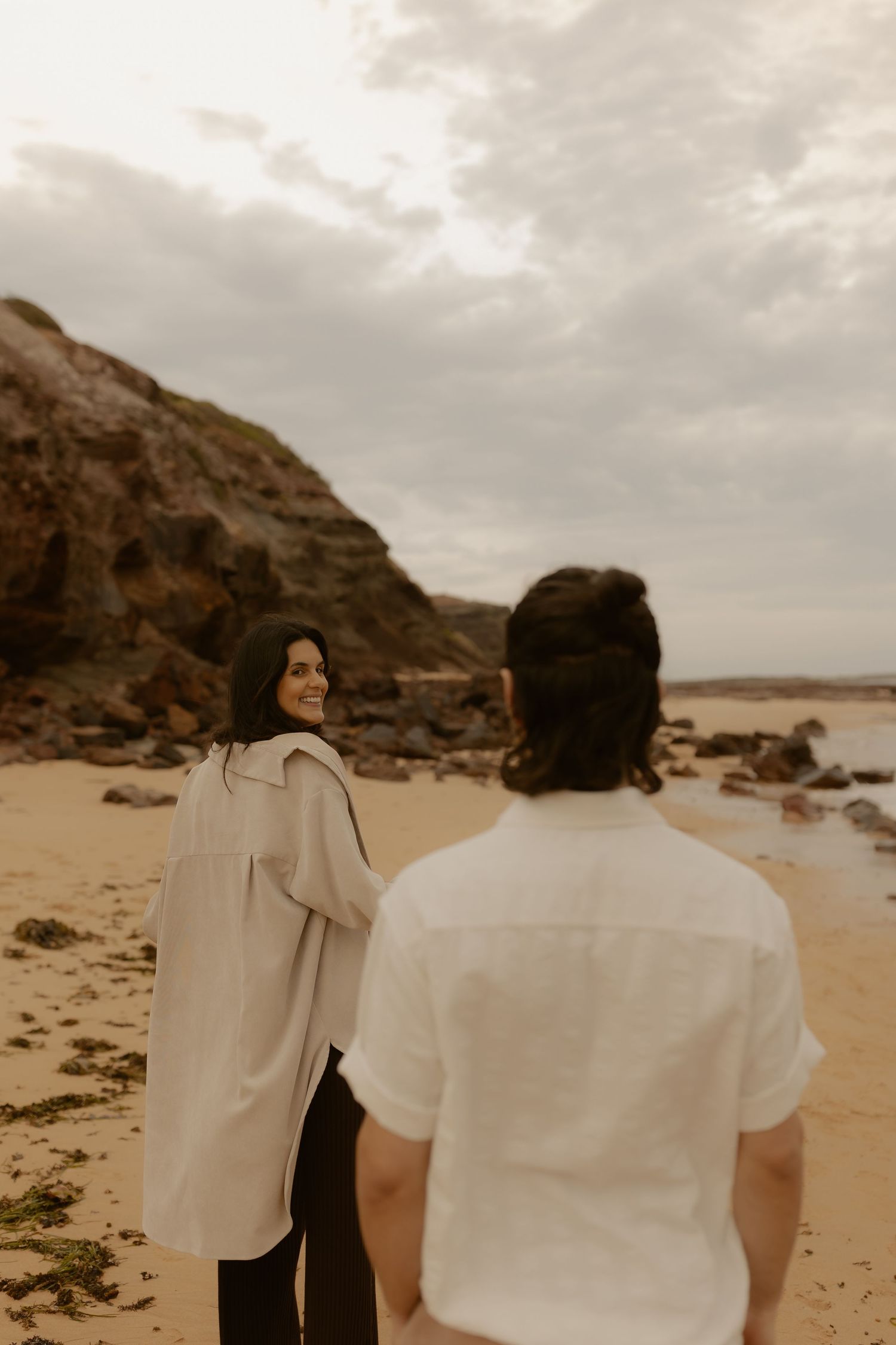 Two figures in white clothing walk along a scenic beach with rocky cliffs and cloudy skies.