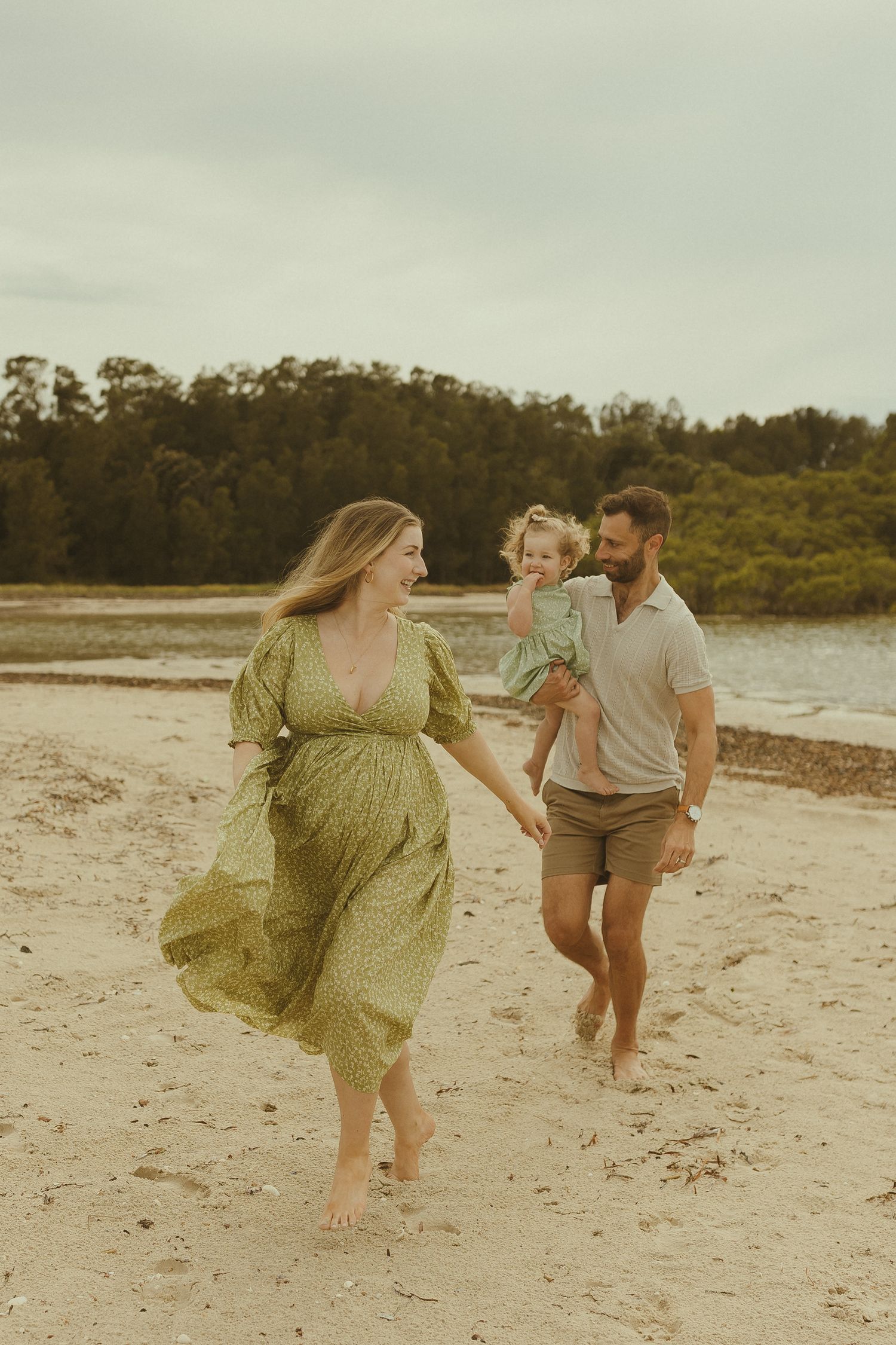 A family plays and runs together on a sandy beach with trees in the background on an overcast day.