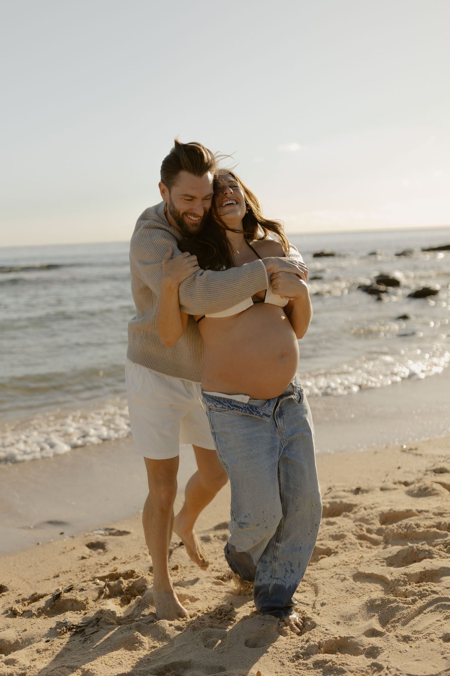 Intimate beach maternity photo of couple embracing on sandy shore at dusk.