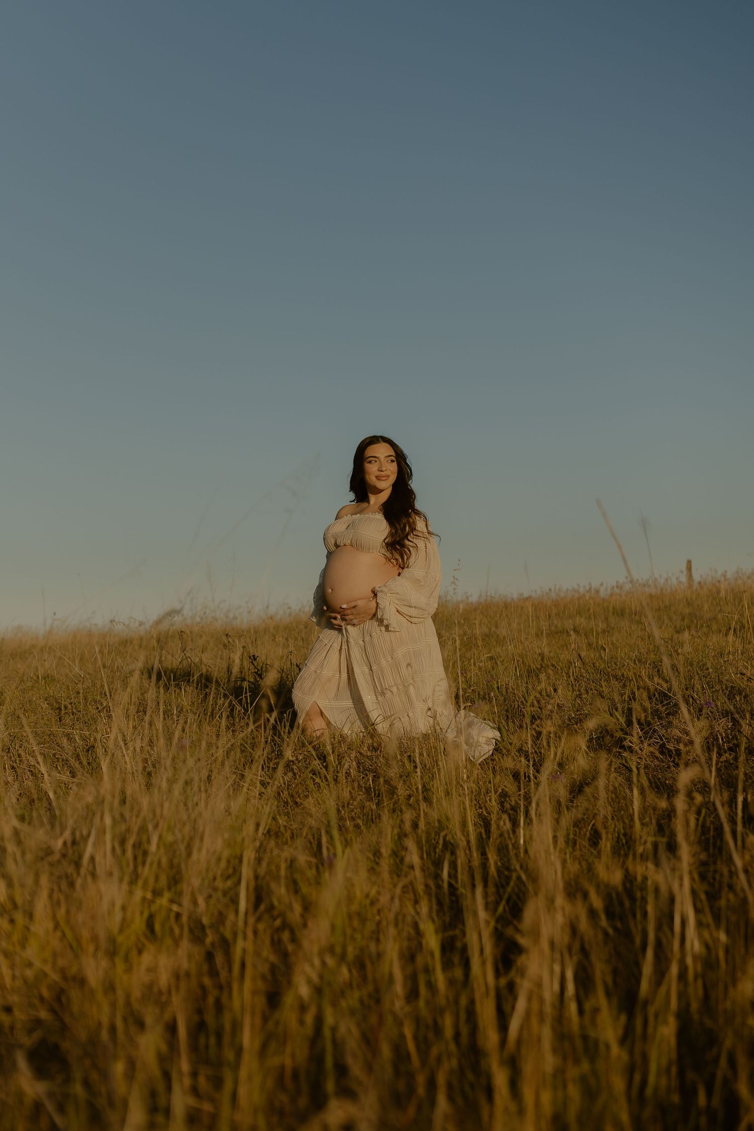 A person in a flowing white dress sits gracefully among tall golden grass in a serene field at sunset.