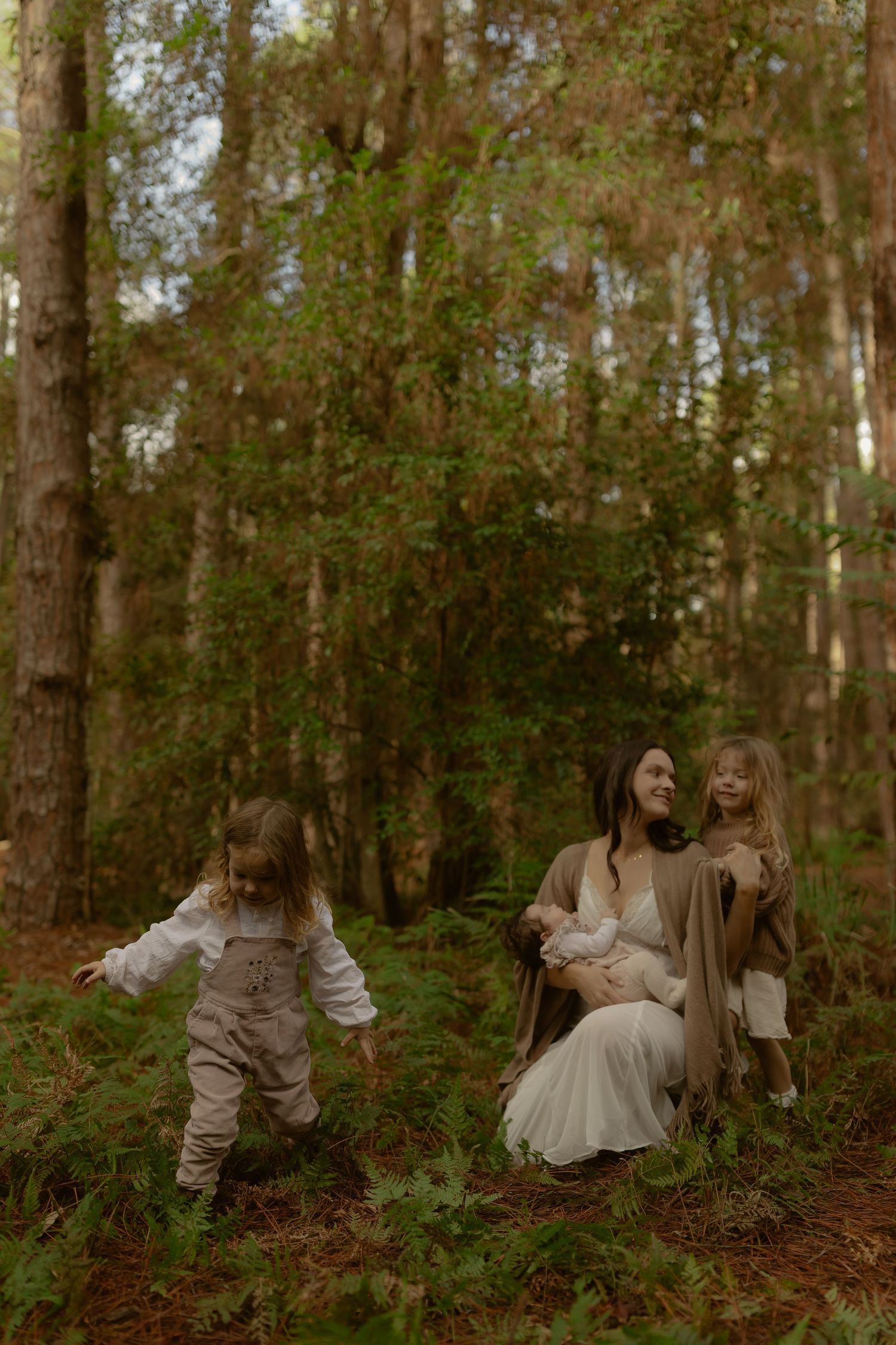 Mother sits in forest with newborn and toddler while young child explores nearby ferns.