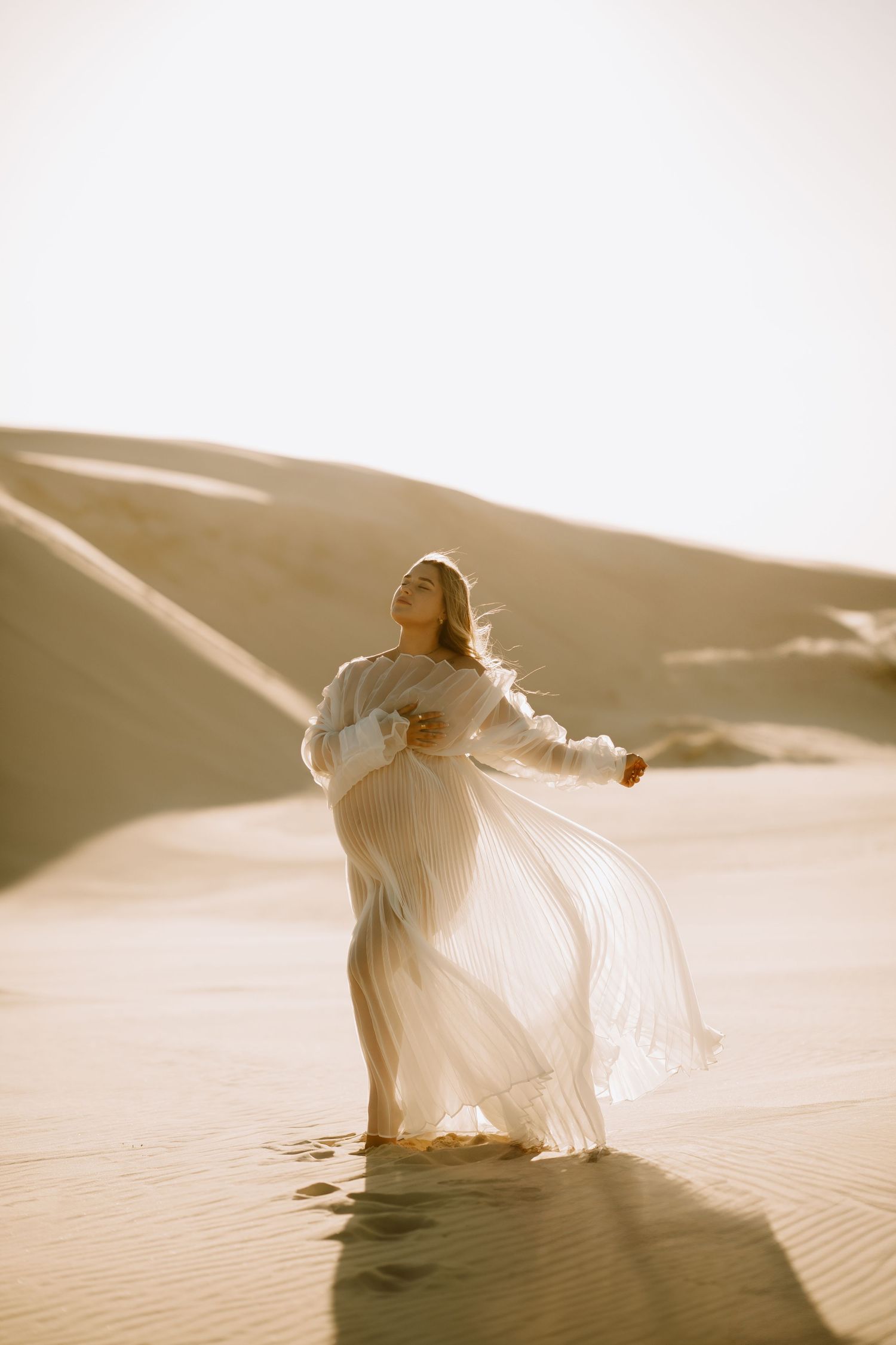 An ethereal maternity scene shows a woman in billowing white dress dancing on sand dunes in golden sunlight.
