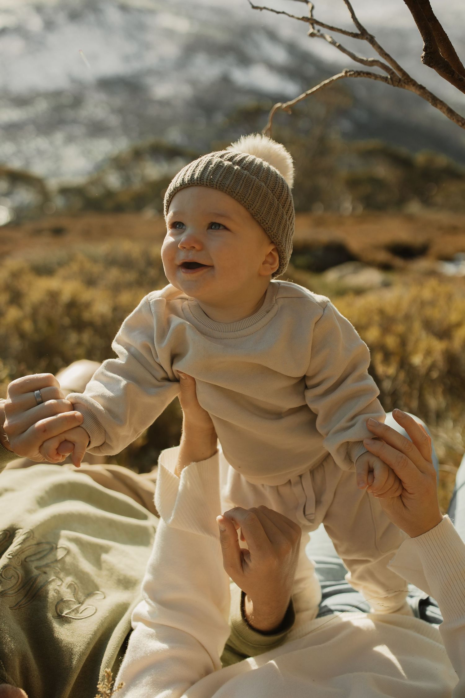 A happy baby wearing a gray knit hat and cream-colored outfit sits outdoors during golden hour.