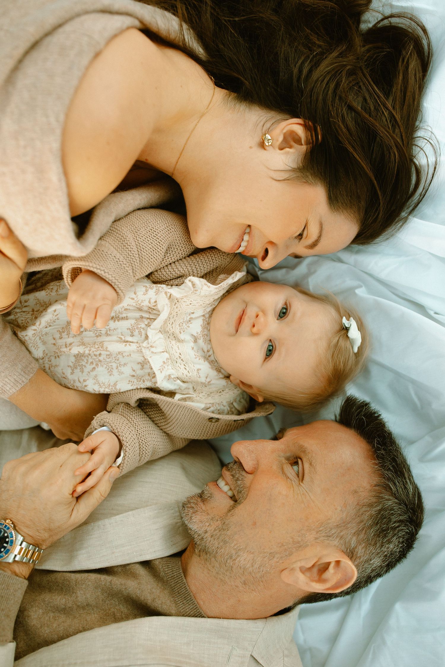 Parents and baby lie together on white blanket during intimate family portrait session.