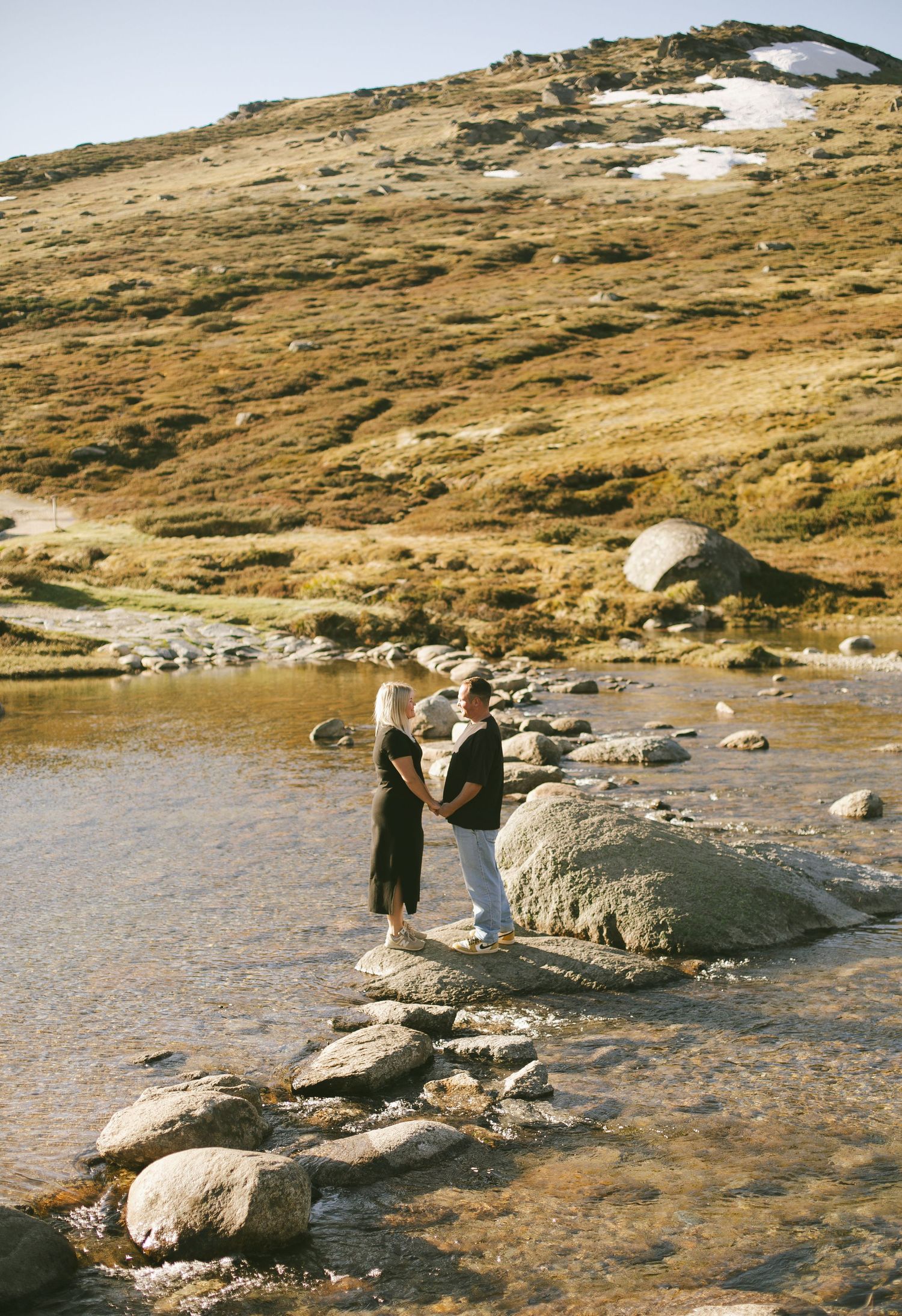 Two figures standing on rocky terrain near a mountain stream with patches of snow visible on distant slopes.