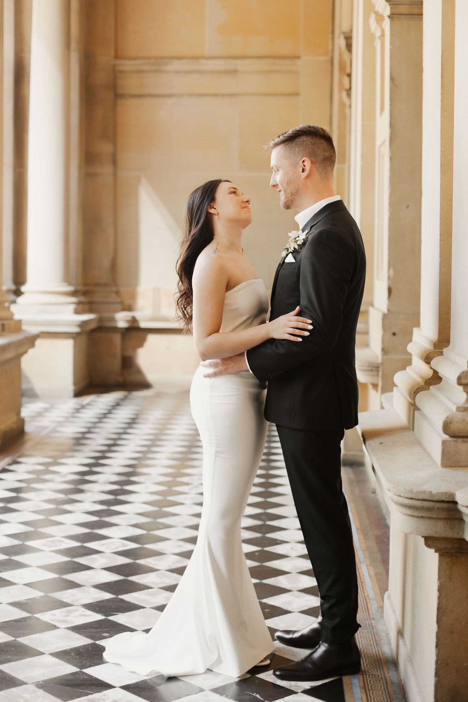 Newlyweds share an intimate moment in a historic building with checkered floor and elegant columns.