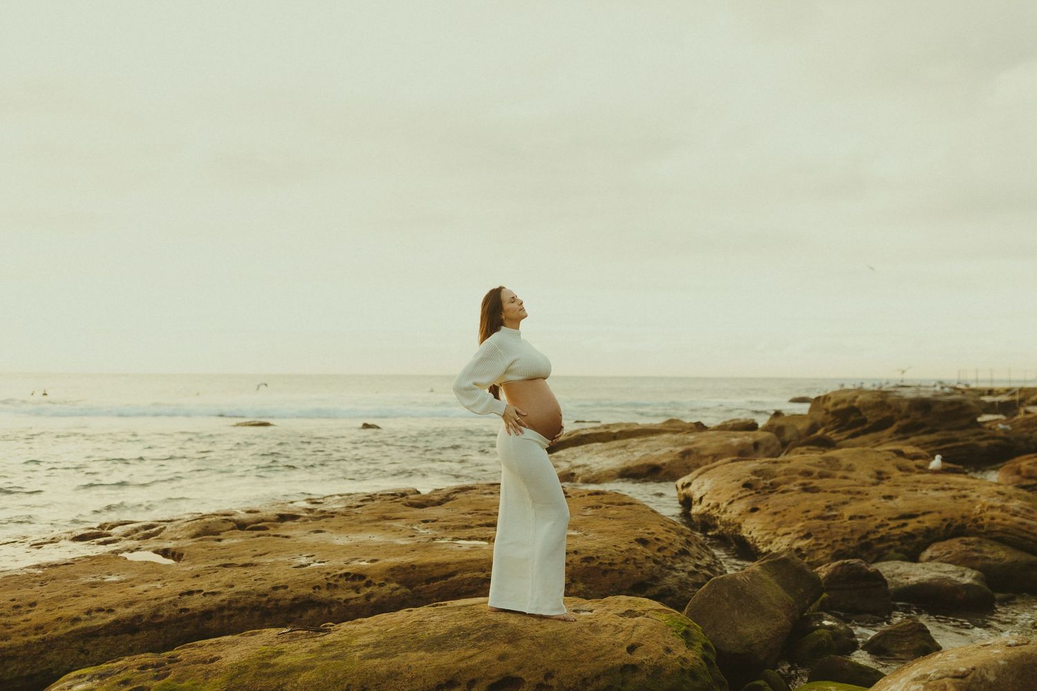 A woman in a flowing white dress stands on coastal rocks overlooking the ocean during golden hour.