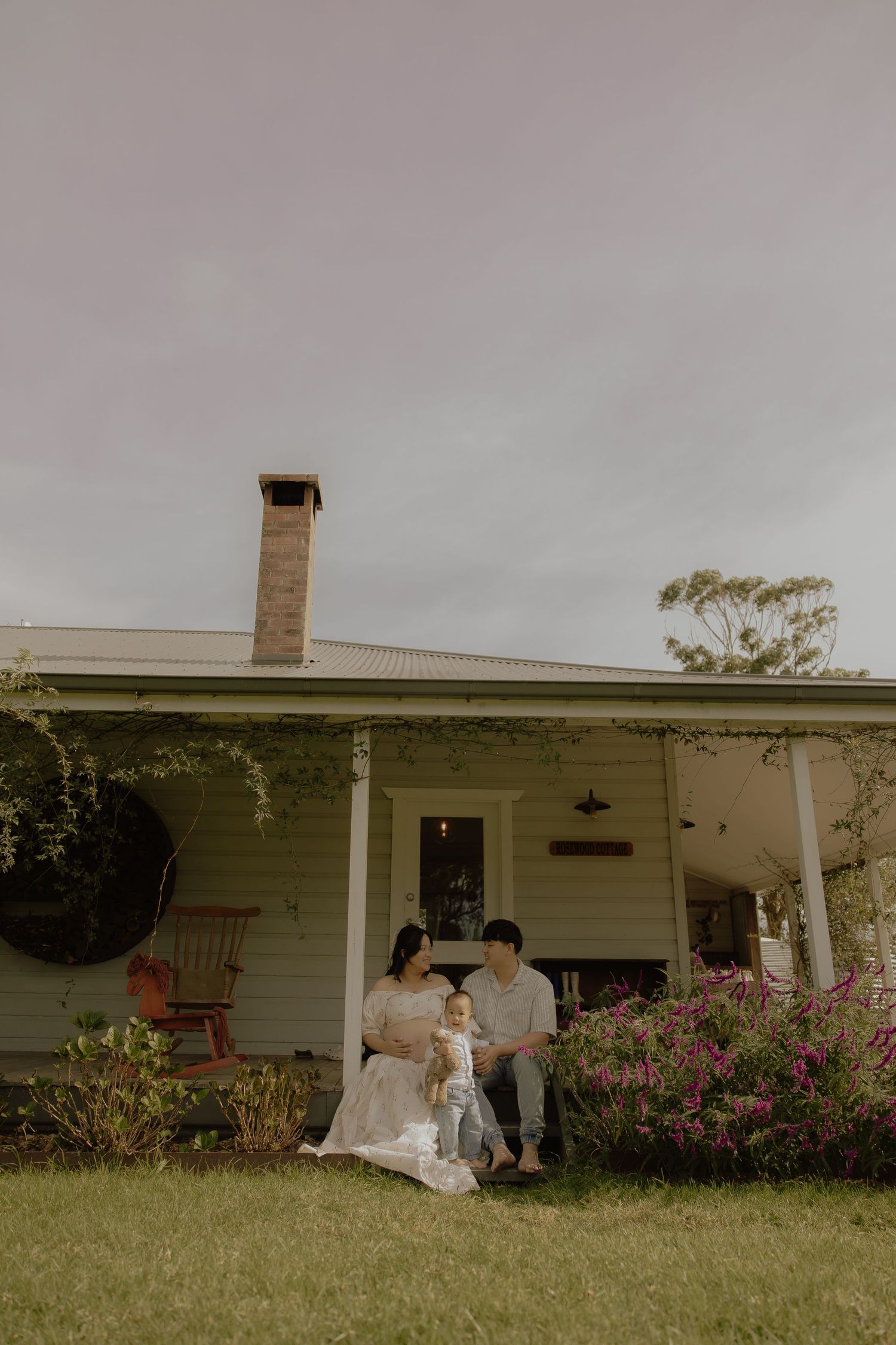 Family with baby sits on lawn in front of charming white cottage farmhouse with pink flowering bushes.