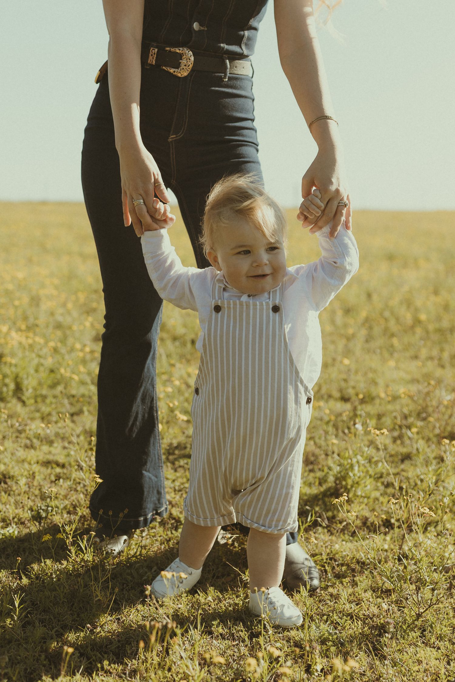 A toddler in striped overalls takes first steps in a sunny field while being supported.