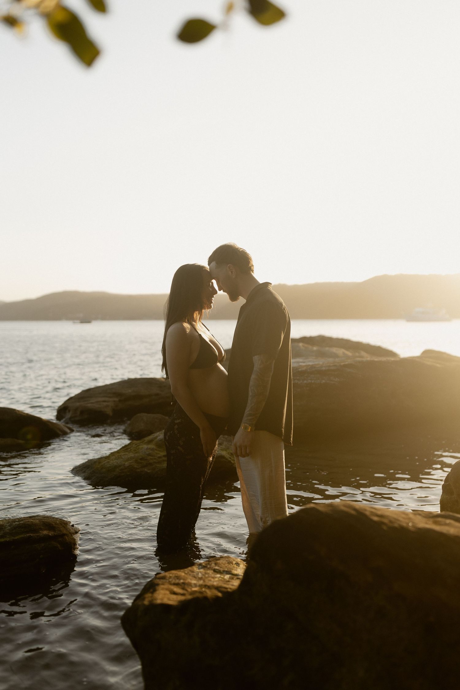 A romantic silhouette of a couple standing on rocky shores at sunset with water and mountains in the background.