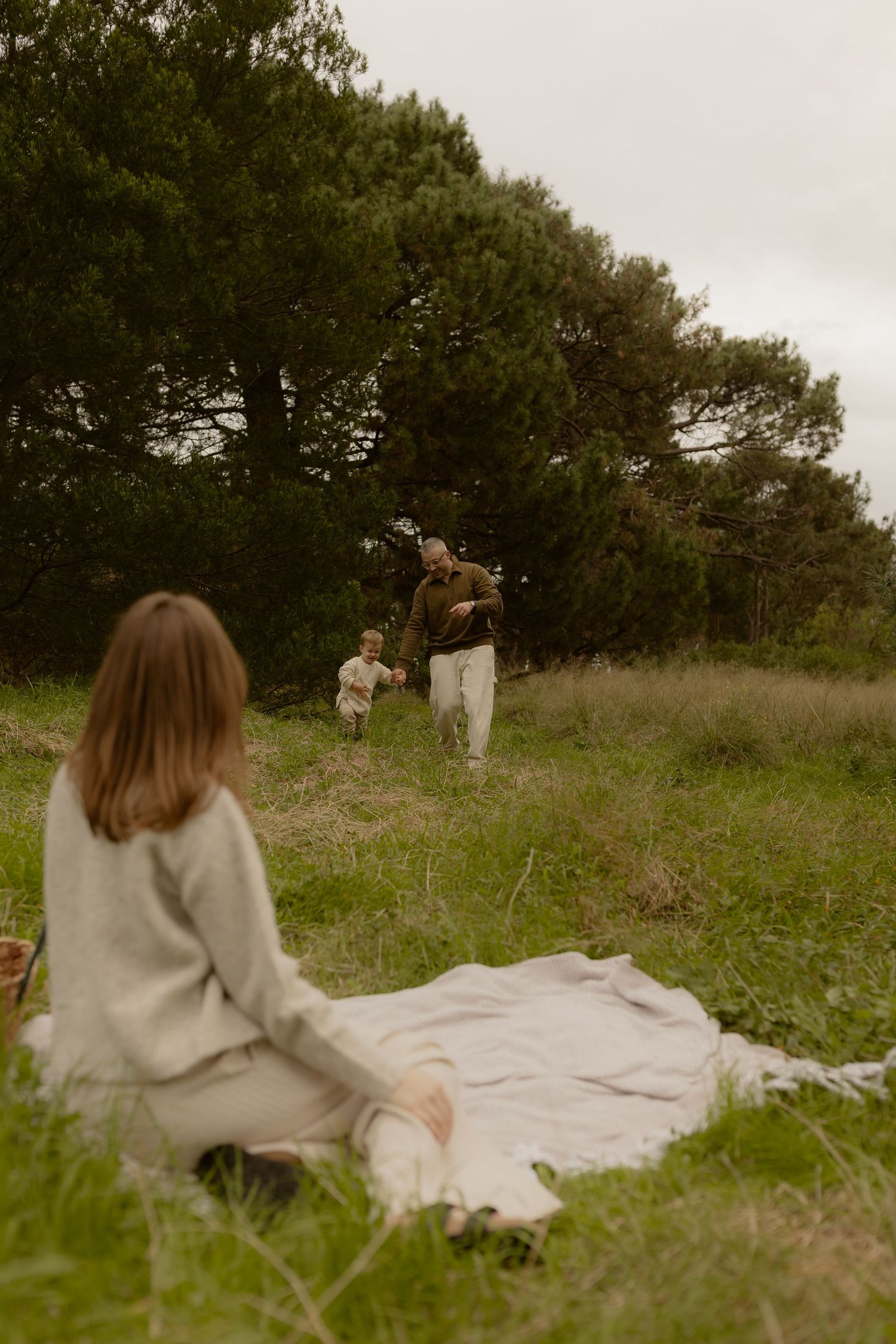 A father plays with his toddler in a green field while a woman sits on a blanket in the foreground.
