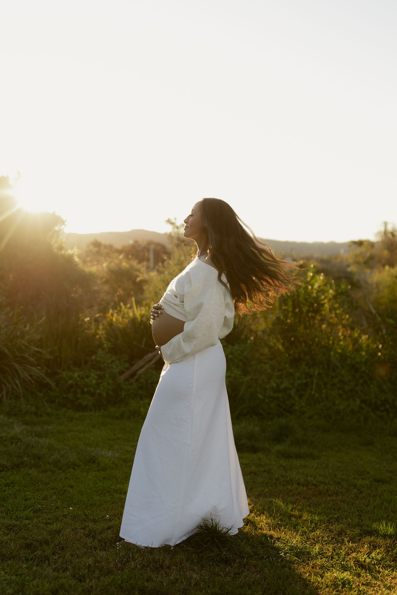 Pregnant woman in white dress standing in golden sunset light outdoors, cradling her baby bump in a serene natural setting.