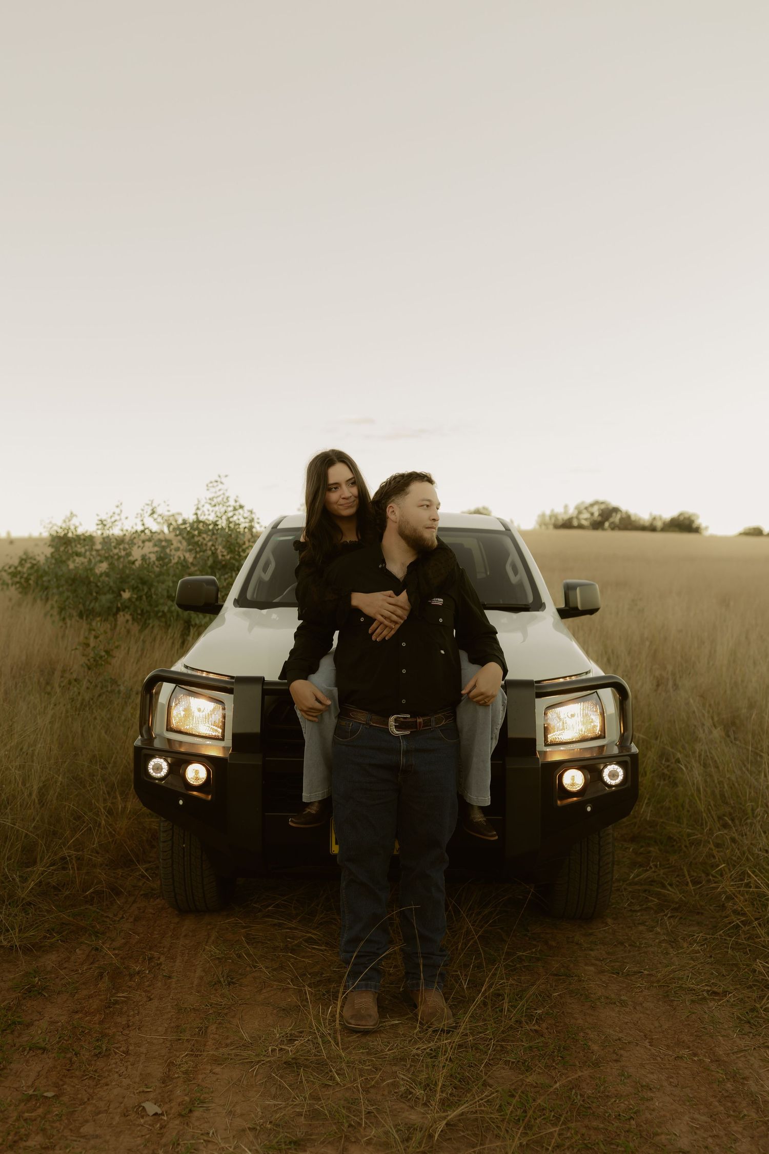 Couple posing in front of a truck on a dirt road surrounded by golden fields at dusk.