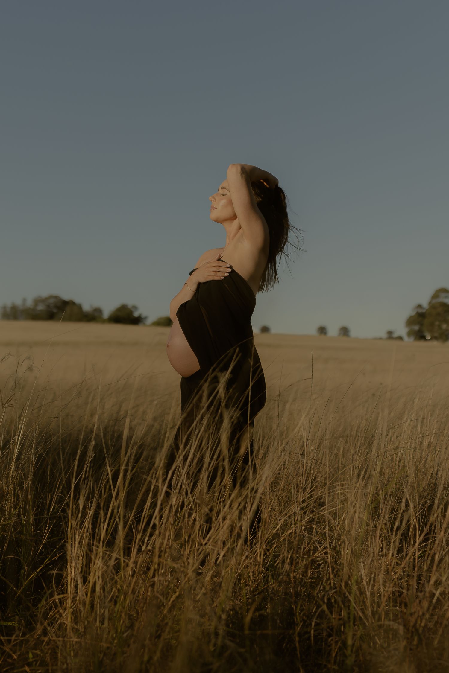Side profile of pregnant woman in black wrap smiling in tall grass field at sunset.