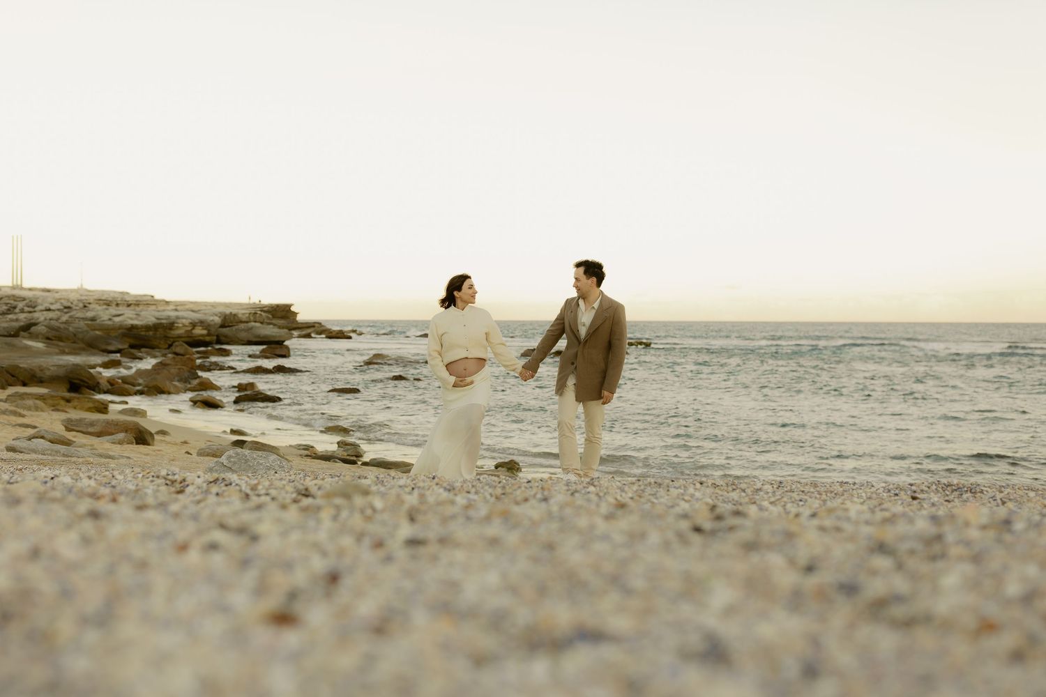 Couple in wedding attire holding hands and walking along a rocky beach at sunset.