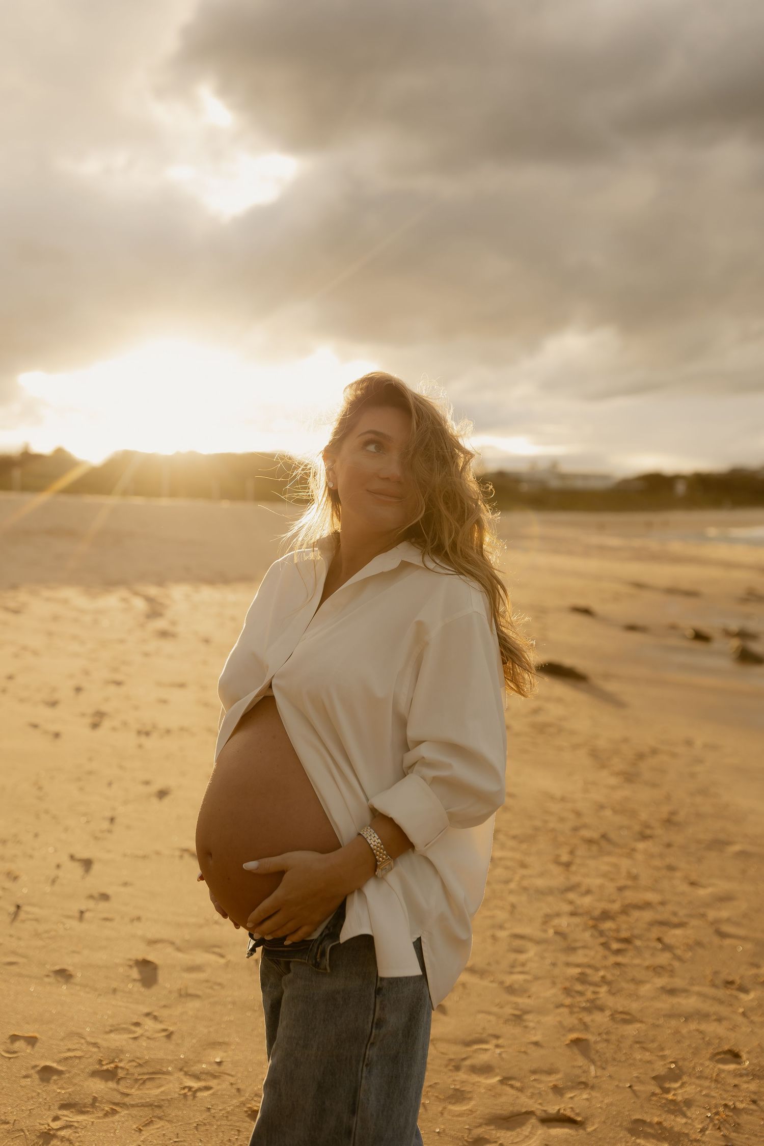 Pregnant woman in white shirt and jeans cradling baby bump on sandy beach at golden sunset.