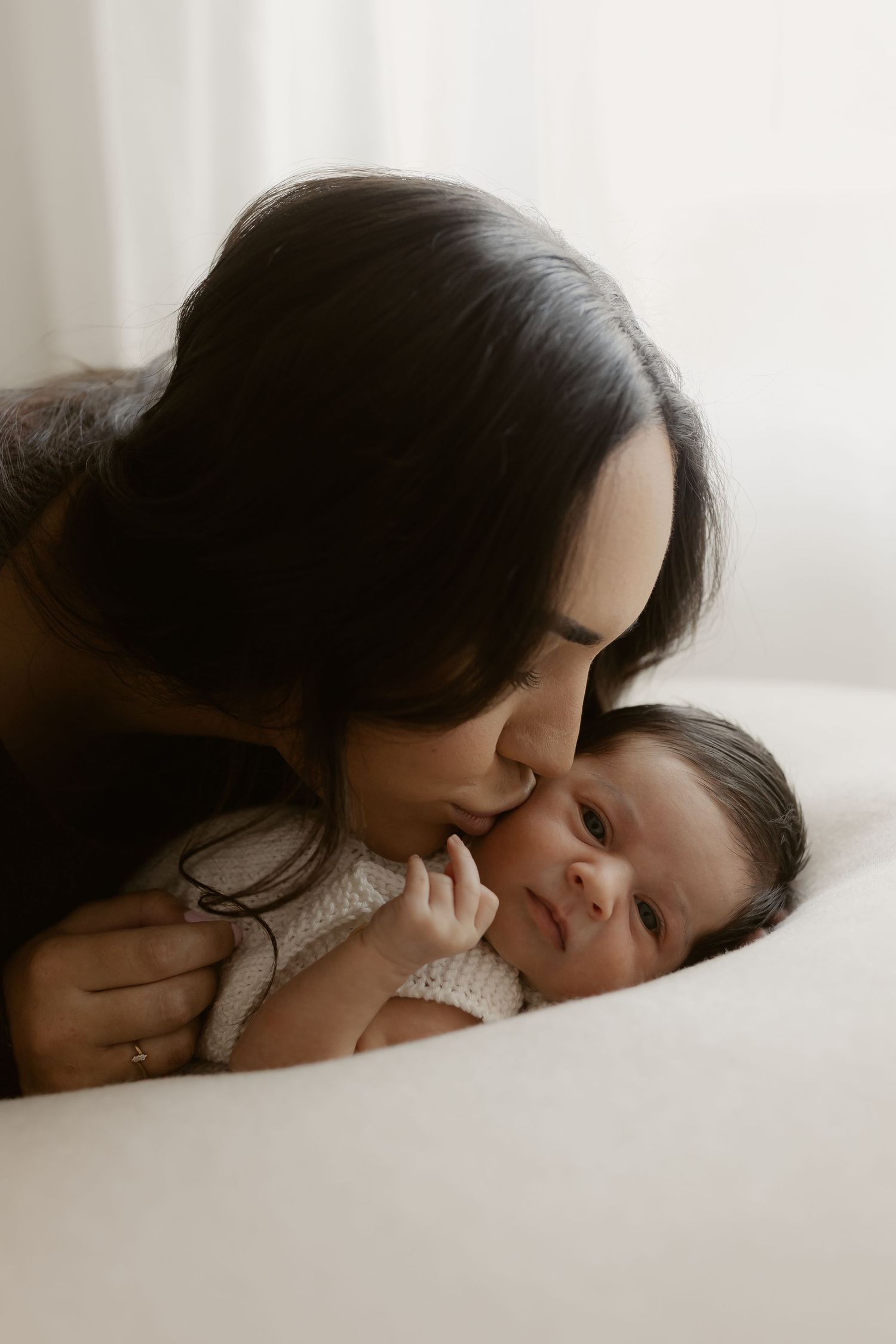 A dark-haired mother tenderly kisses her newborn baby lying on a white surface in a soft, natural light setting.