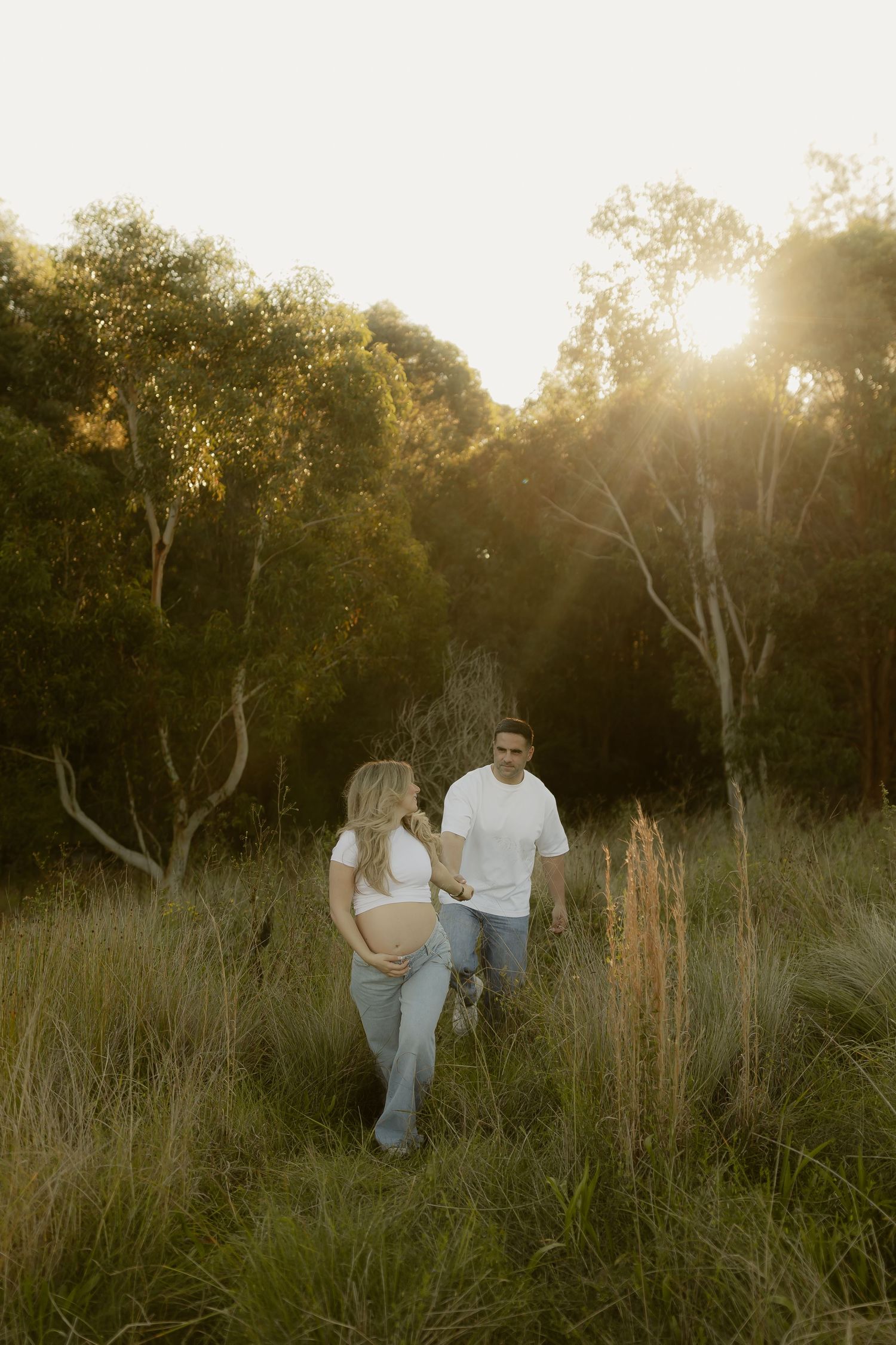 Pregnant couple walking through golden sunlit field surrounded by tall grass and eucalyptus trees at sunset.