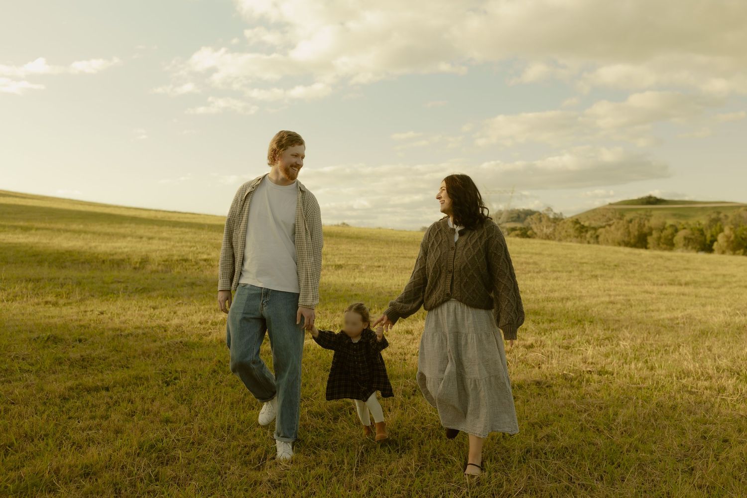 A happy family of three walks hand-in-hand through a lush green meadow under a cloudy sky.