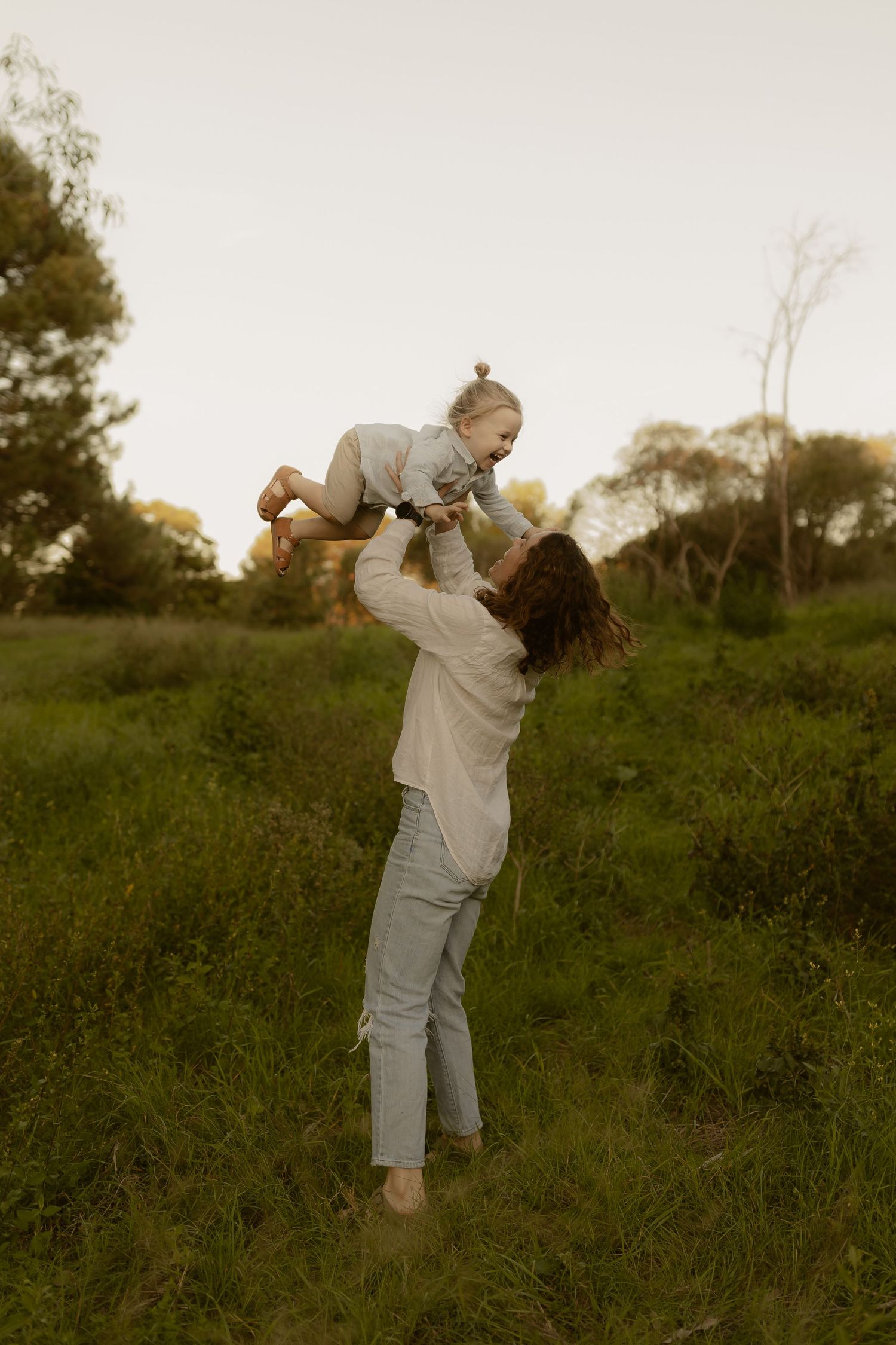 A couple in light colored clothing embraces and dances together in a grassy field at sunset.