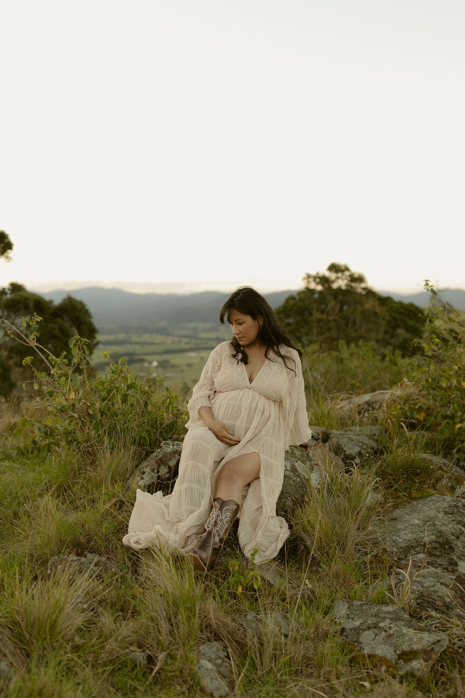 Pregnant woman in white lace dress and boots sitting on rocks in a scenic hillside landscape at dusk.